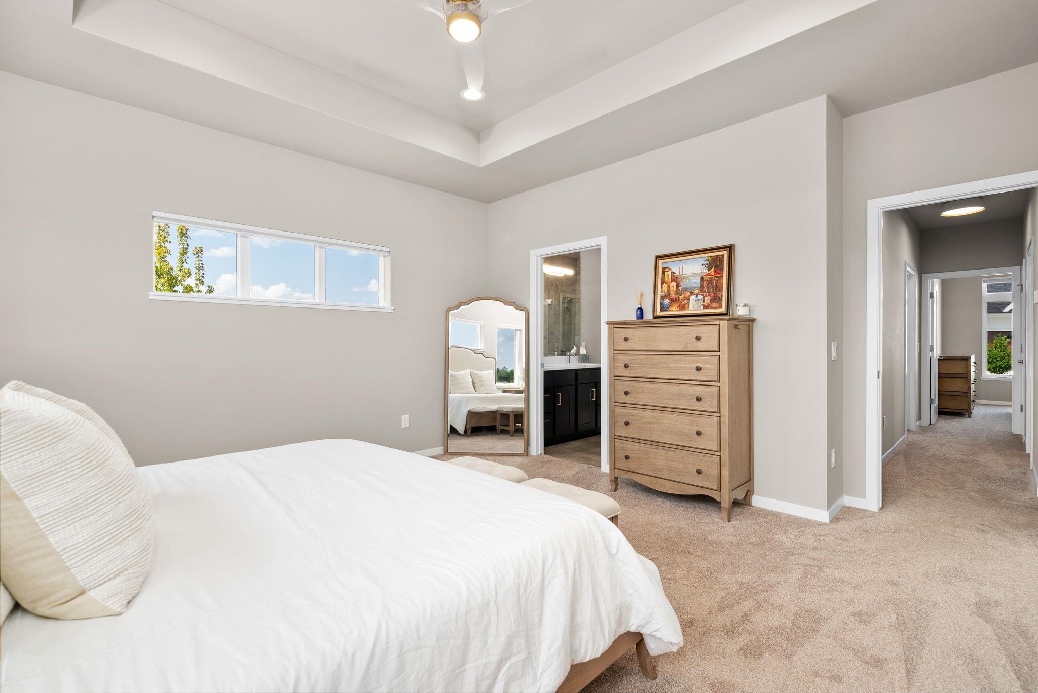 Furnished bedroom with a tray ceiling, horizontal window, wooden chest, and doorway to an ensuite bathroom.