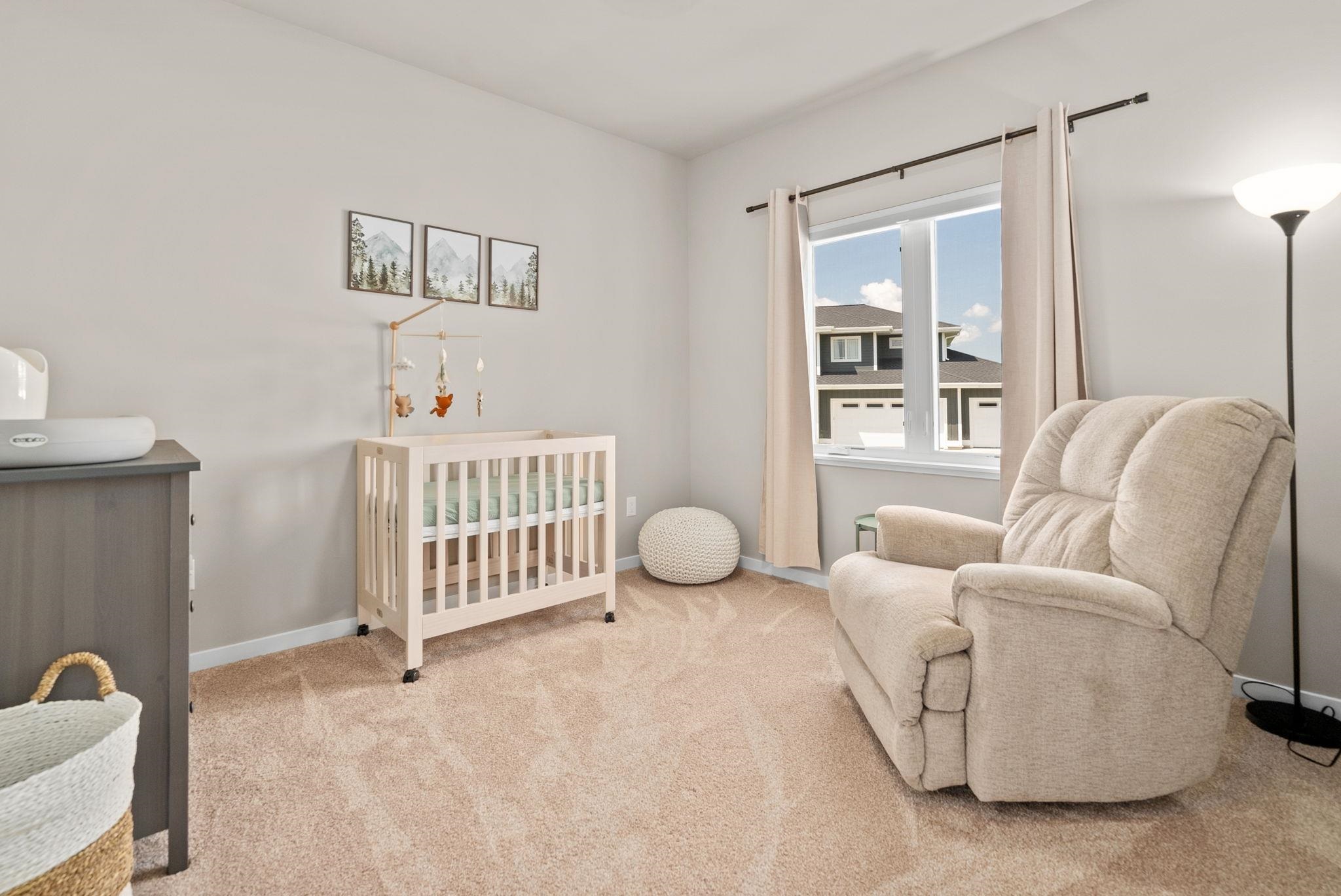 Furnished nursery with a light wood crib, plush recliner, and window overlooking a neighboring home.