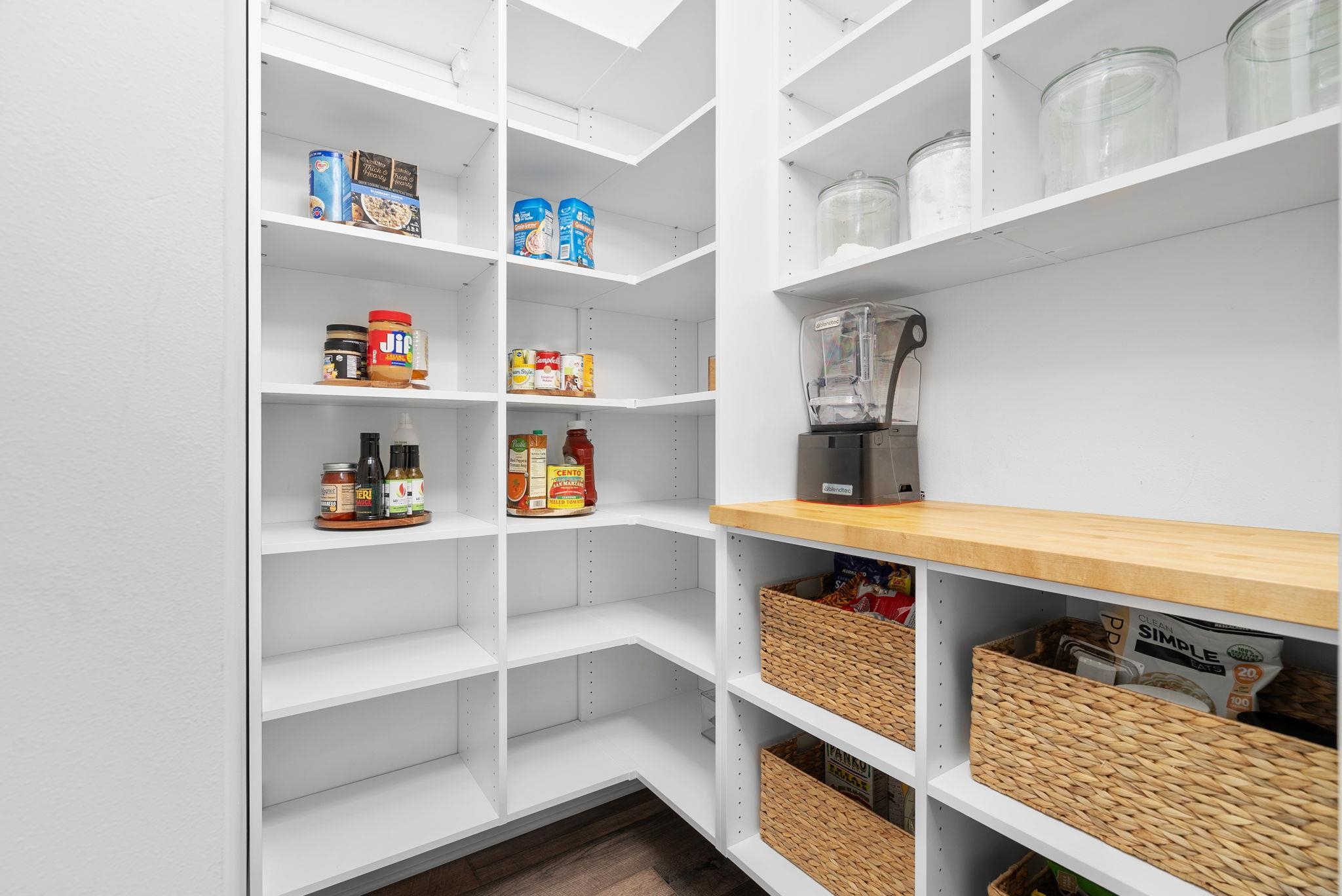 Walk-in pantry with white built-in shelves, a butcher-block counter, blender, and woven baskets.