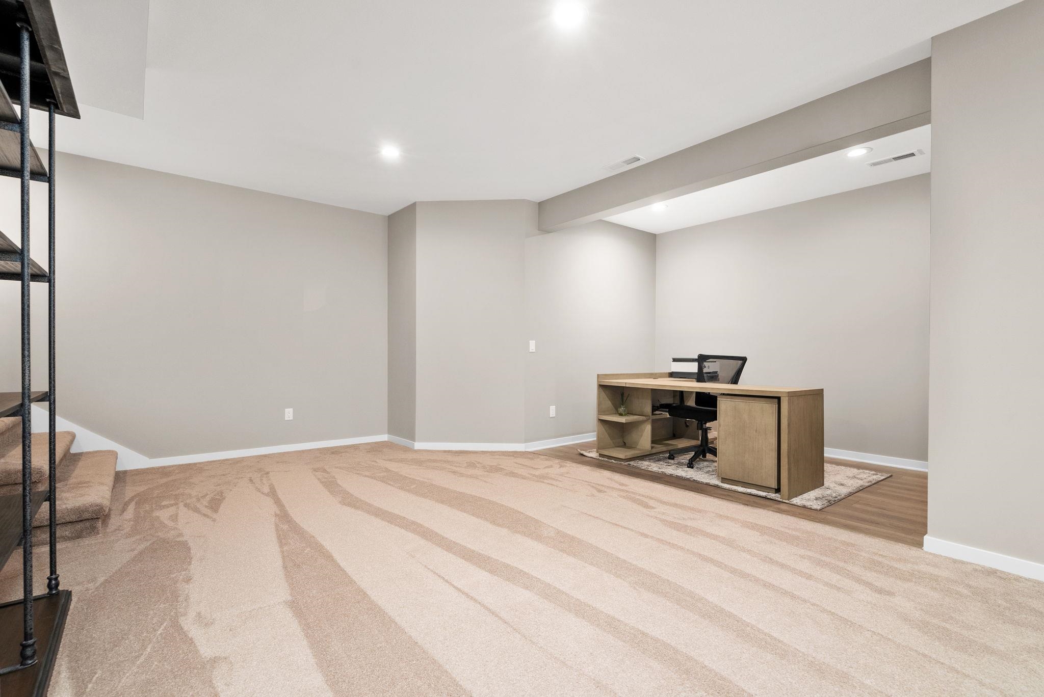 Basement office with a wooden desk and chair, patterned carpet, and carpeted stairs.