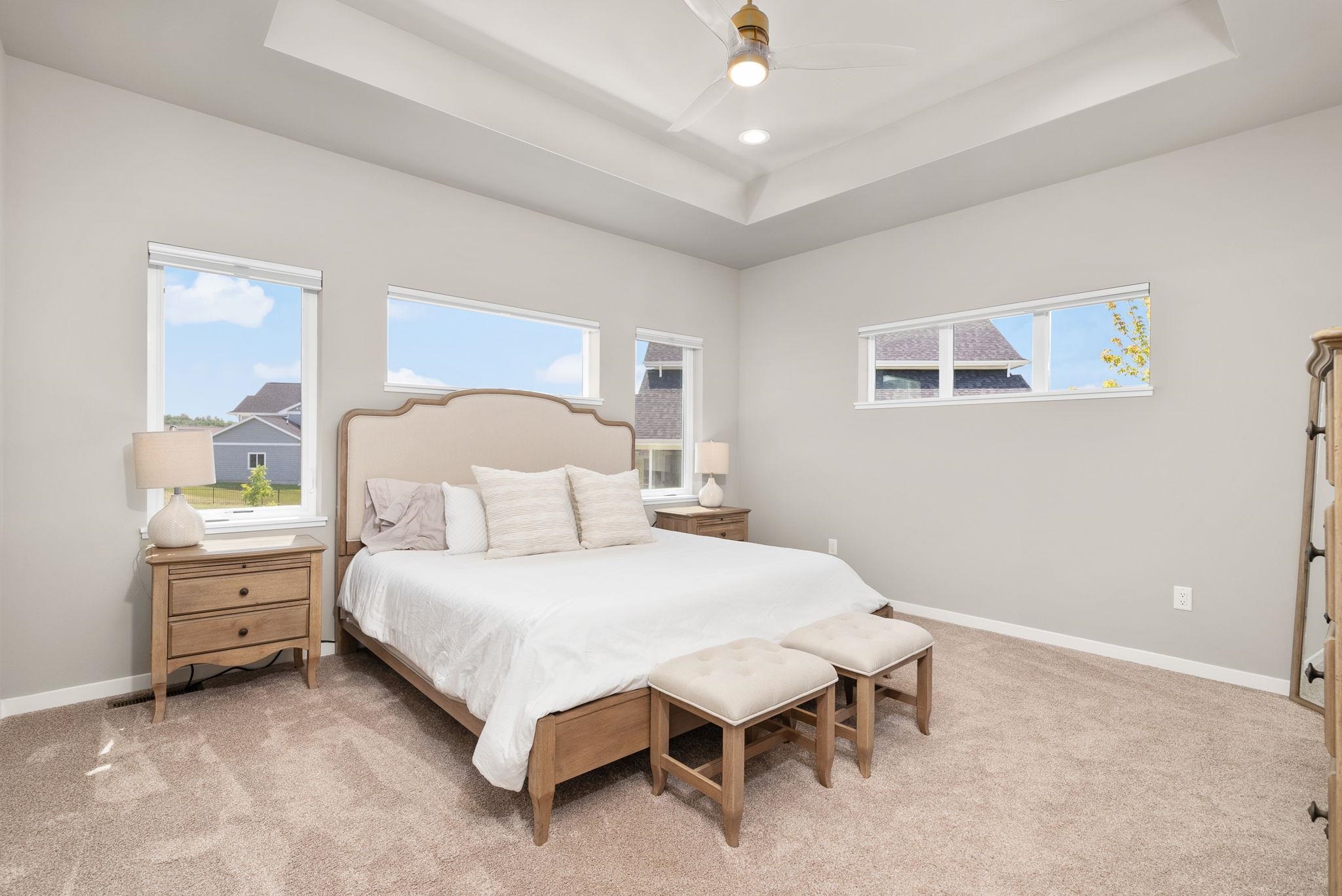 Staged bedroom with a large bed, two cushioned benches, tray ceiling, and several windows.