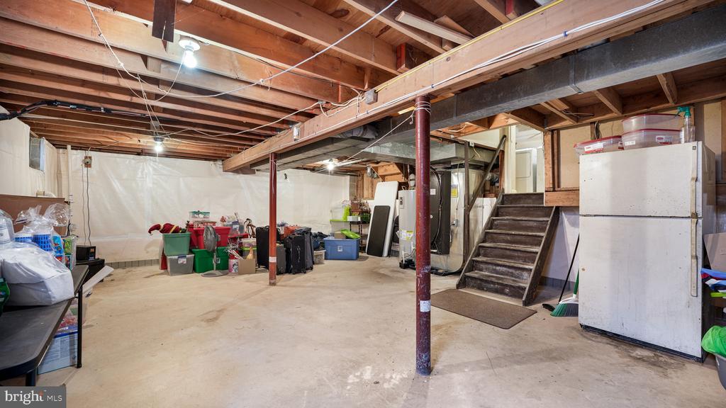 Unfinished basement with exposed ceiling joists, support posts, and stairs beside a refrigerator.