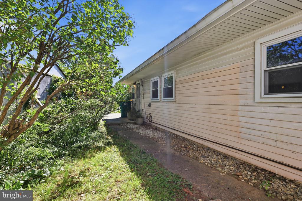 Side of a light-colored house beside a concrete walkway and mature leafy landscaping.