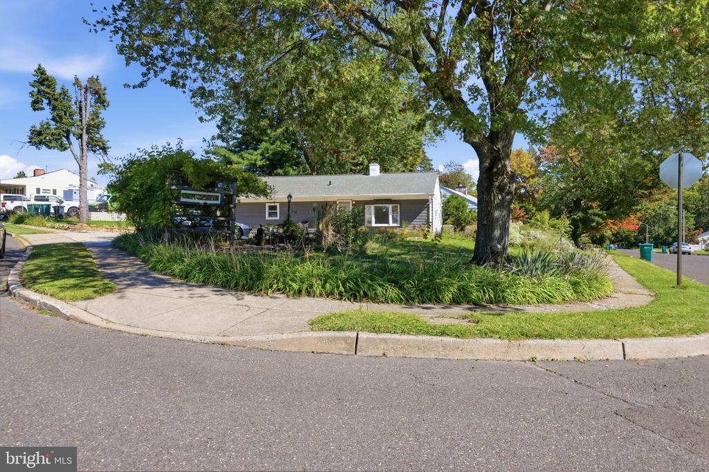 Single-story house on a corner lot beneath a large tree, with landscaped beds and a sidewalk.