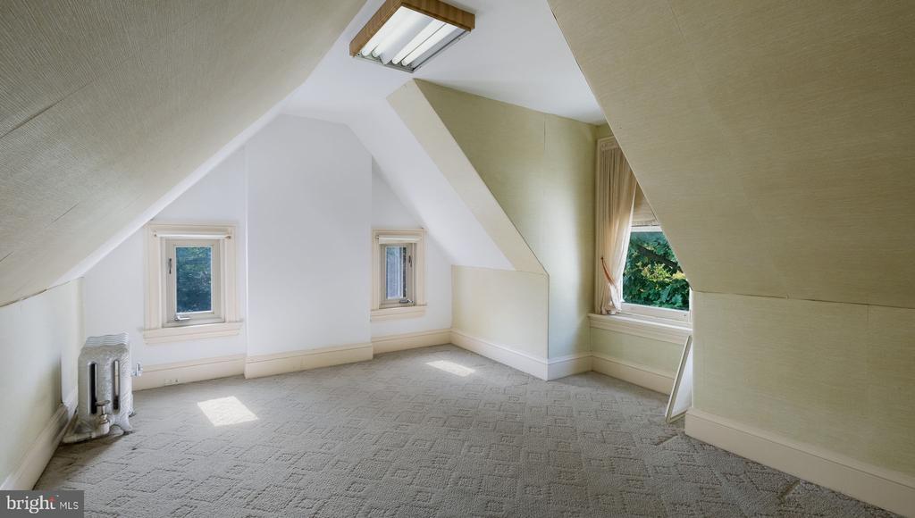 Empty finished attic room with sloped ceilings, beige carpet, three windows, and a radiator.