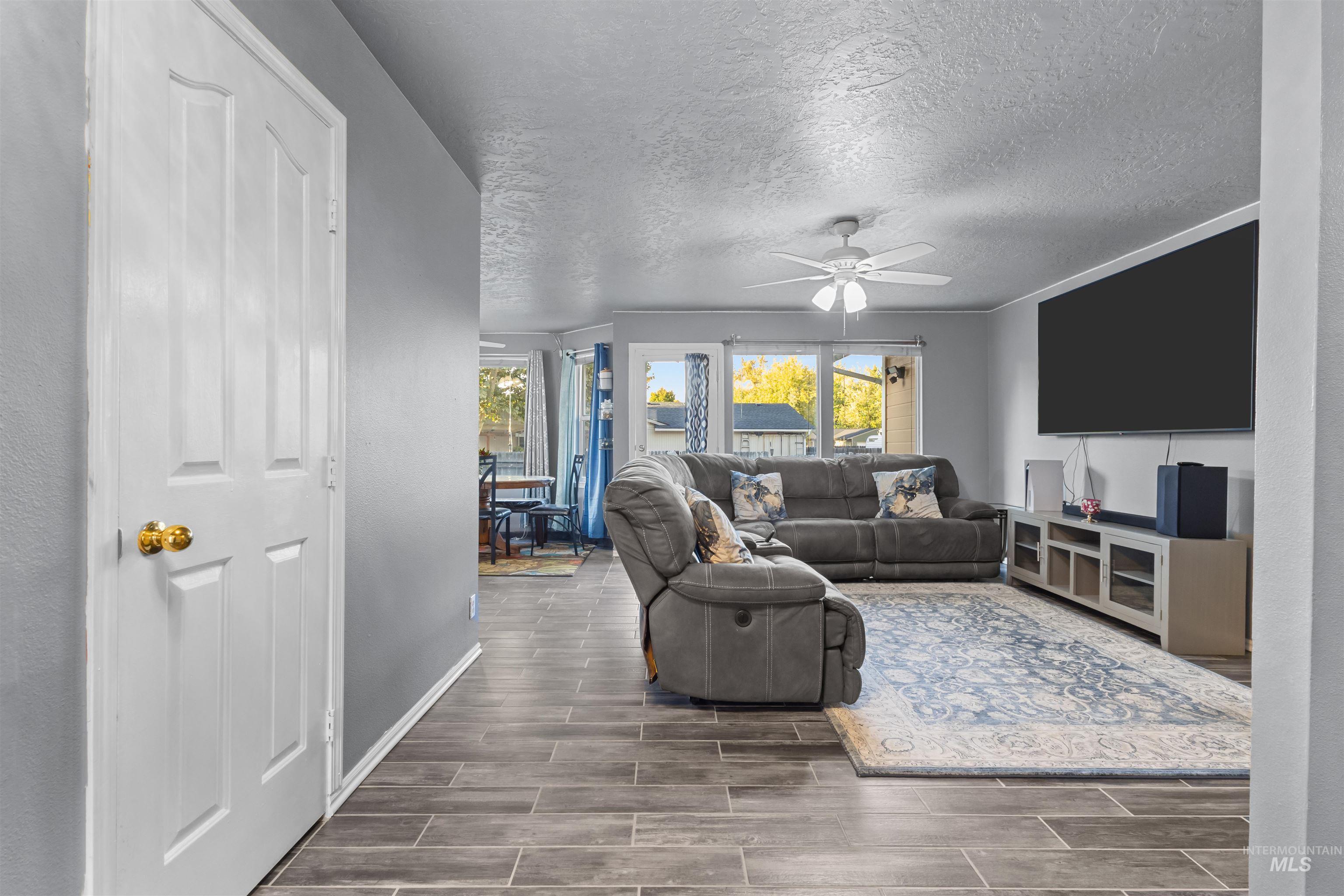 Furnished living room with gray reclining sofas, a wall-mounted TV, and windows overlooking autumn foliage.