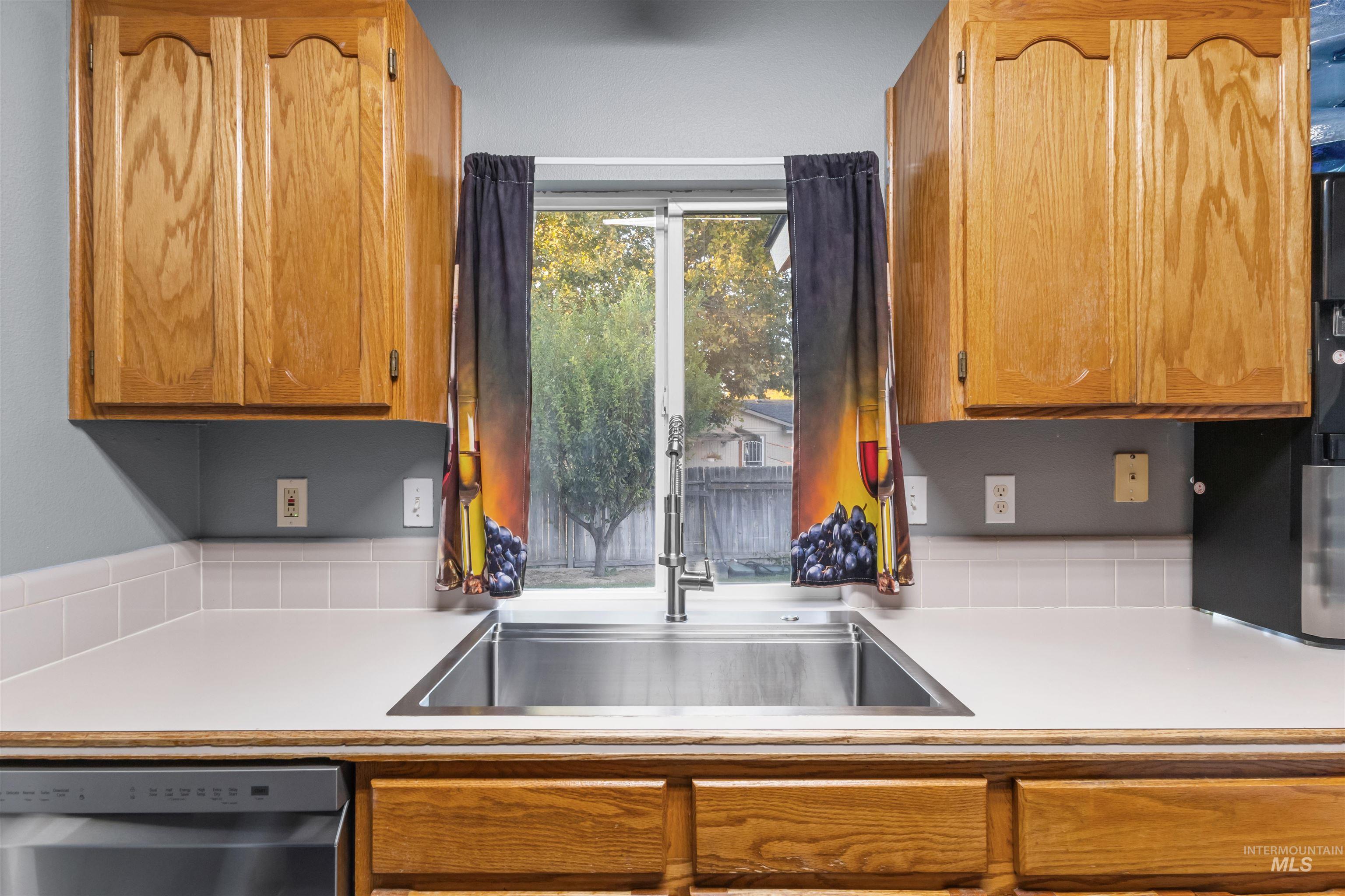 Kitchen sink beneath a window, framed by honey-colored wood cabinets and fruit-patterned curtains.