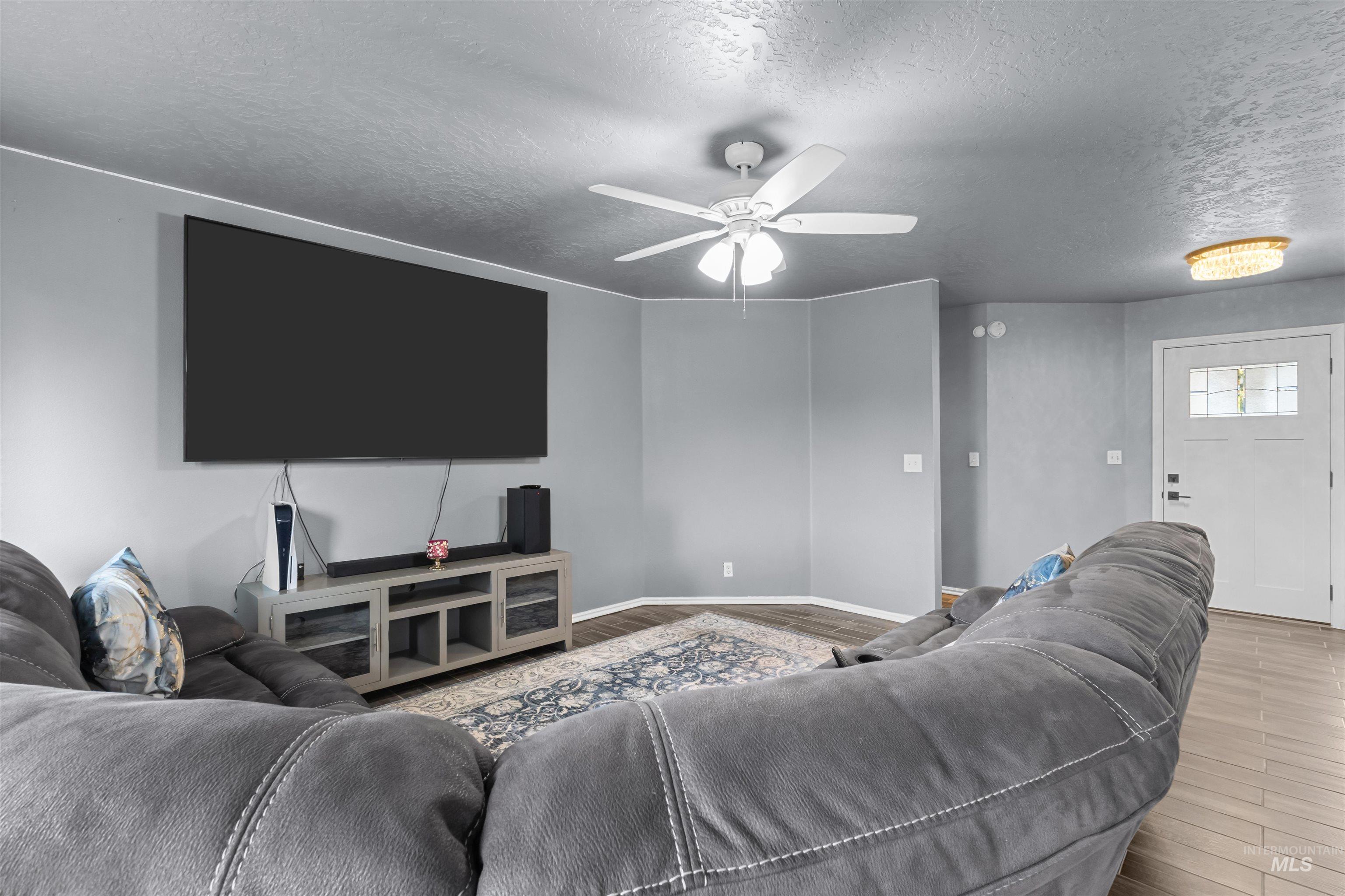 Furnished living room with a gray sectional, large wall-mounted TV, and ceiling fan.