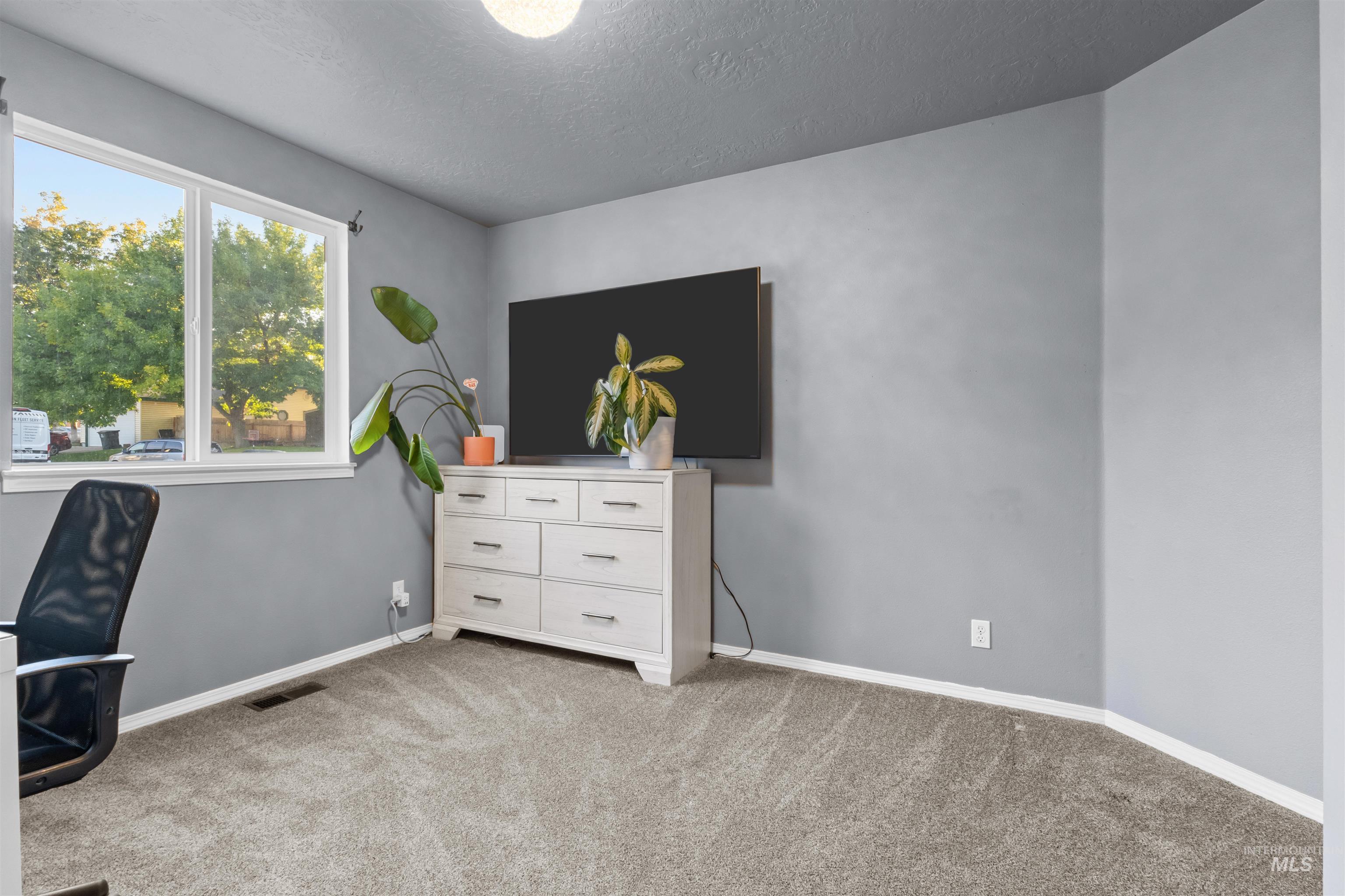 Furnished home office with gray carpet, a large window, and a white dresser beneath a wall-mounted television.
