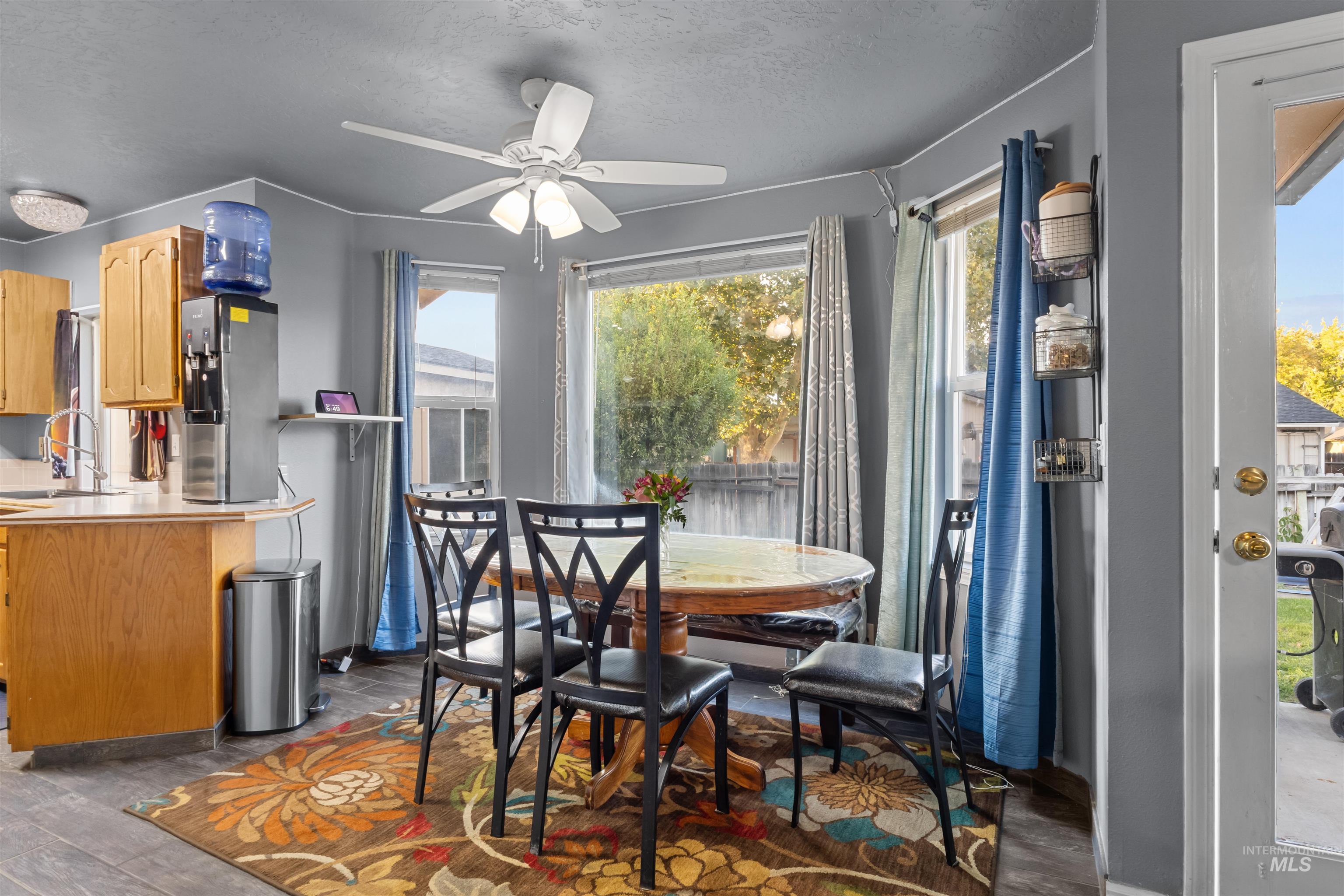 Dining area with a round wooden table, black chairs, and windows overlooking a leafy backyard.