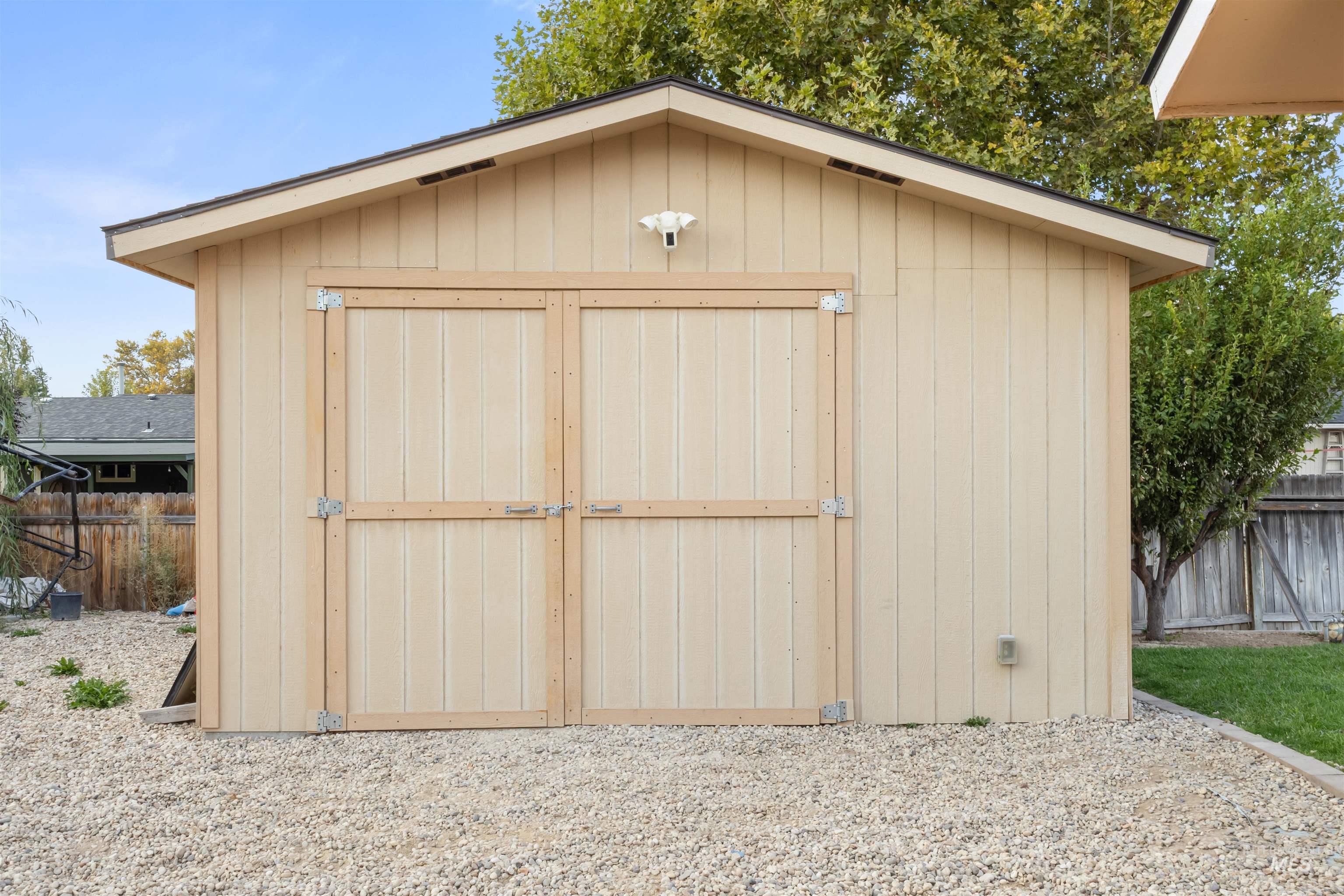 Beige backyard shed with wide double doors and a gabled roof, surrounded by gravel and trees.
