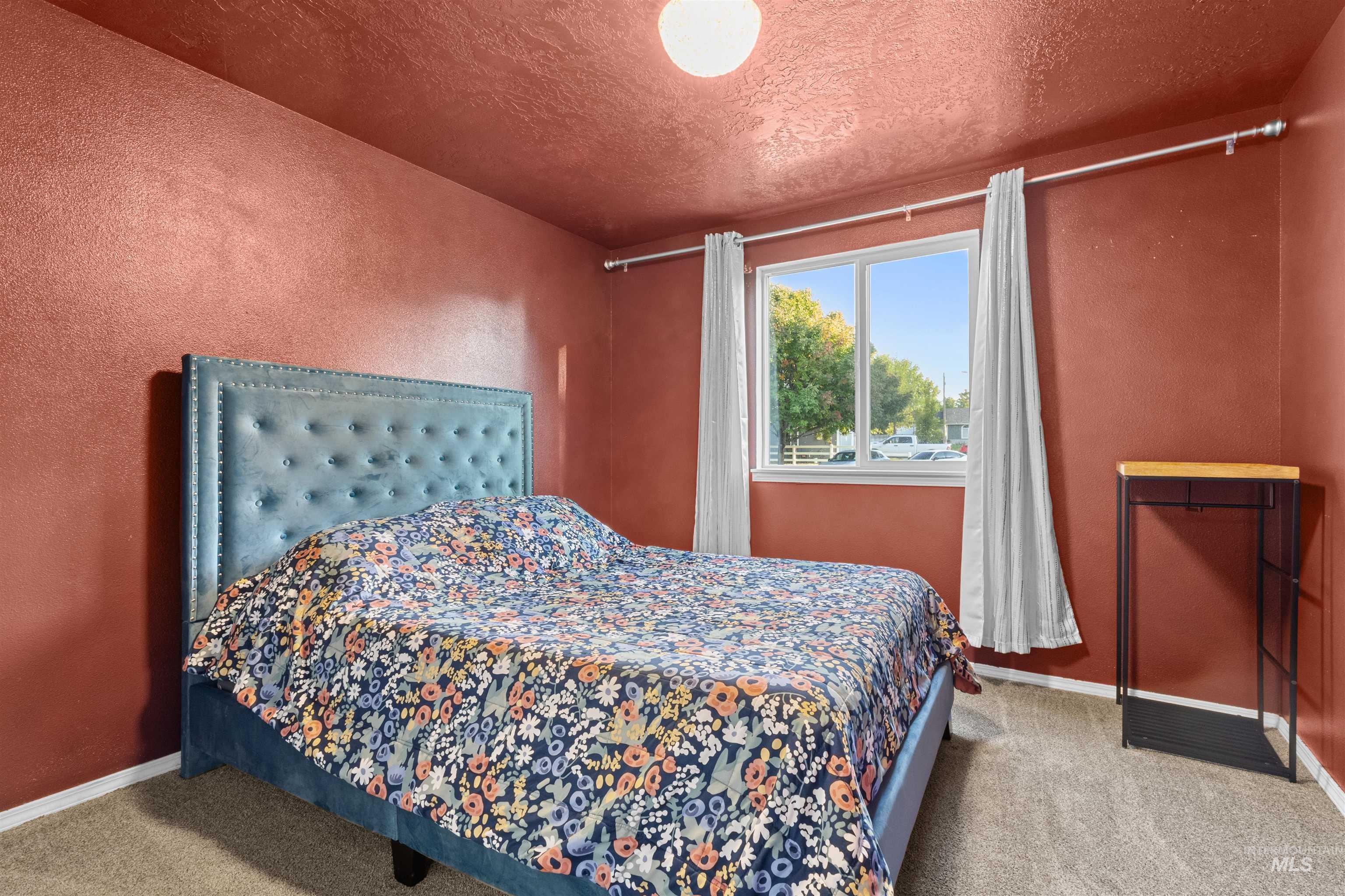 Bedroom with terracotta walls, a blue upholstered bed, floral bedding, and a curtained street-facing window.