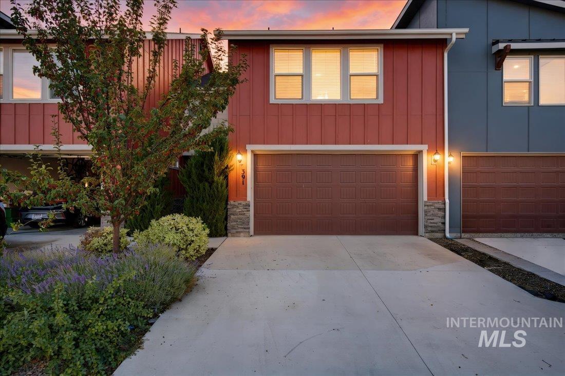 Attached home with red siding, a double garage, landscaped shrubs, and a pink dusk sky.