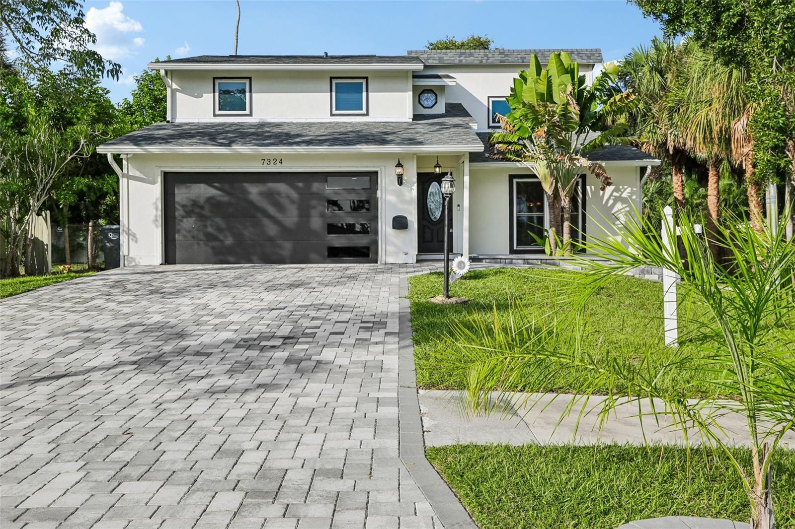 Two-story white house with a dark garage, broad paver driveway, and tropical landscaping.