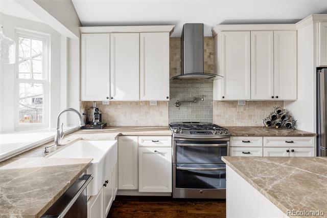 Bright kitchen with white cabinets, stone counters, and a stainless-steel range beneath a chimney hood.