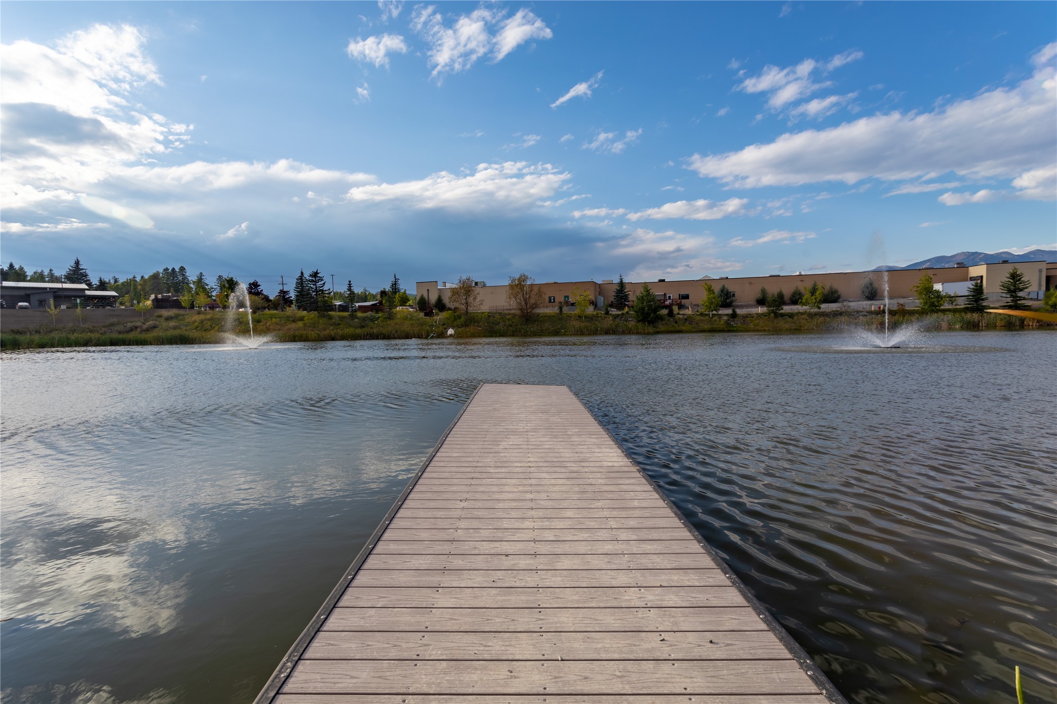 Wooden dock extends into a pond with fountains, landscaped grounds, and distant mountains.
