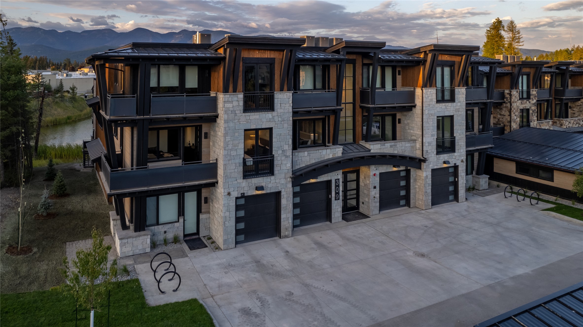 Modern stone-and-wood townhouses with balconies and garage doors beside a waterway, with mountains behind.