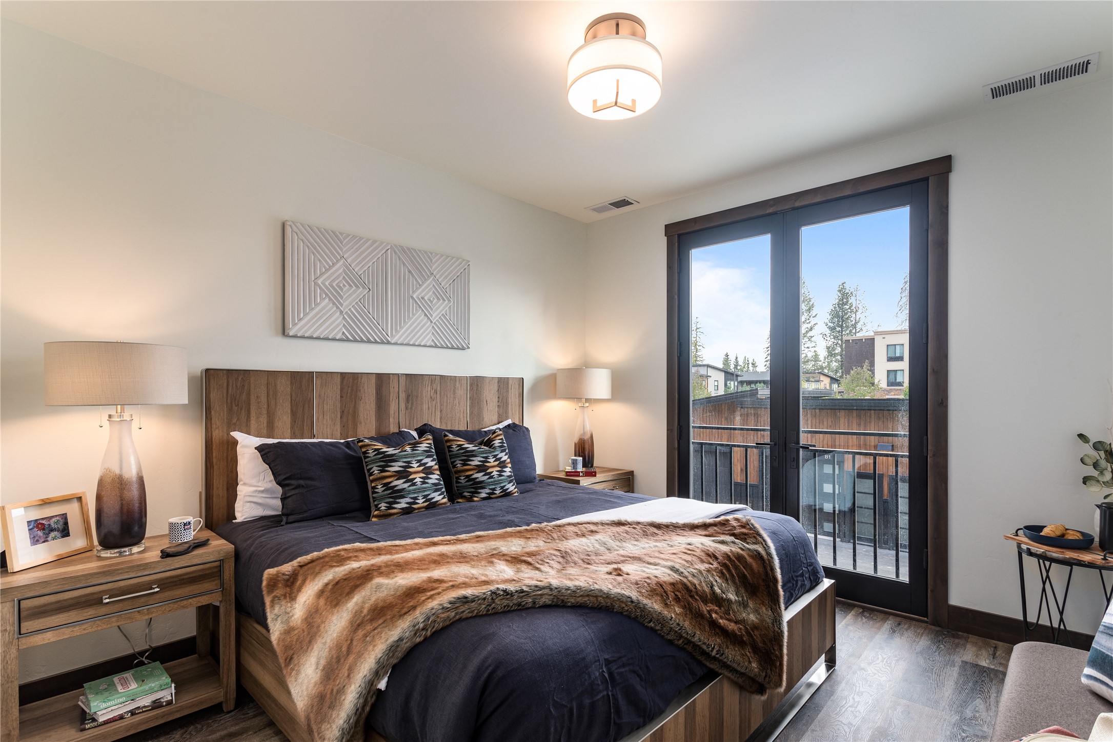 Furnished bedroom with navy bedding and dark-framed balcony doors overlooking nearby buildings and trees.