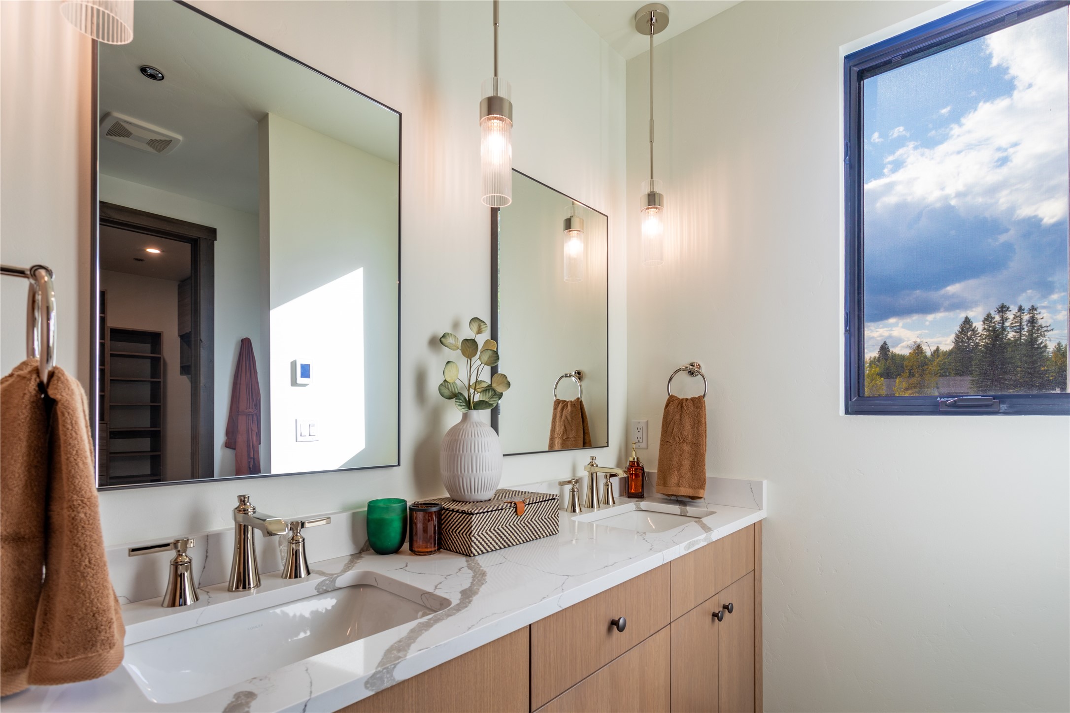 Bathroom double vanity with veined stone counters, wood cabinetry, pendant lights, and a window overlooking trees.