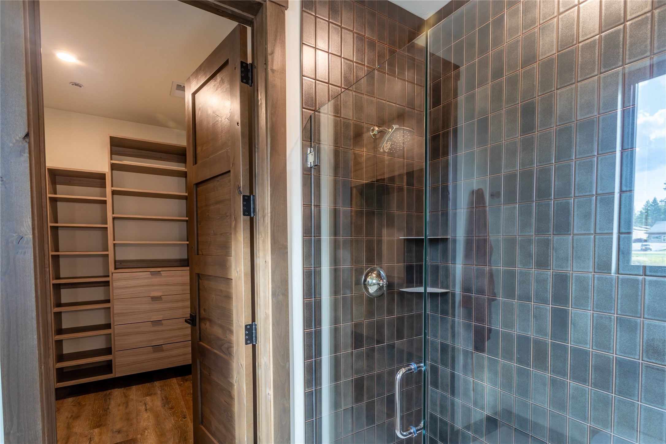 Tiled glass shower beside a walk-in closet with built-in wooden shelves and drawers.