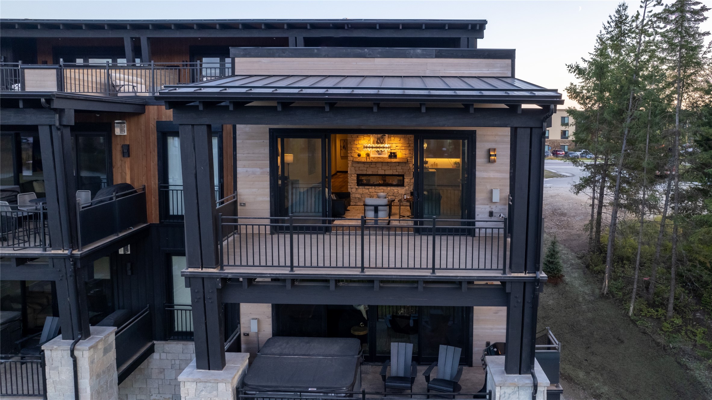 Modern home exterior with a warmly lit fireplace lounge on the upper balcony and a hot tub below.