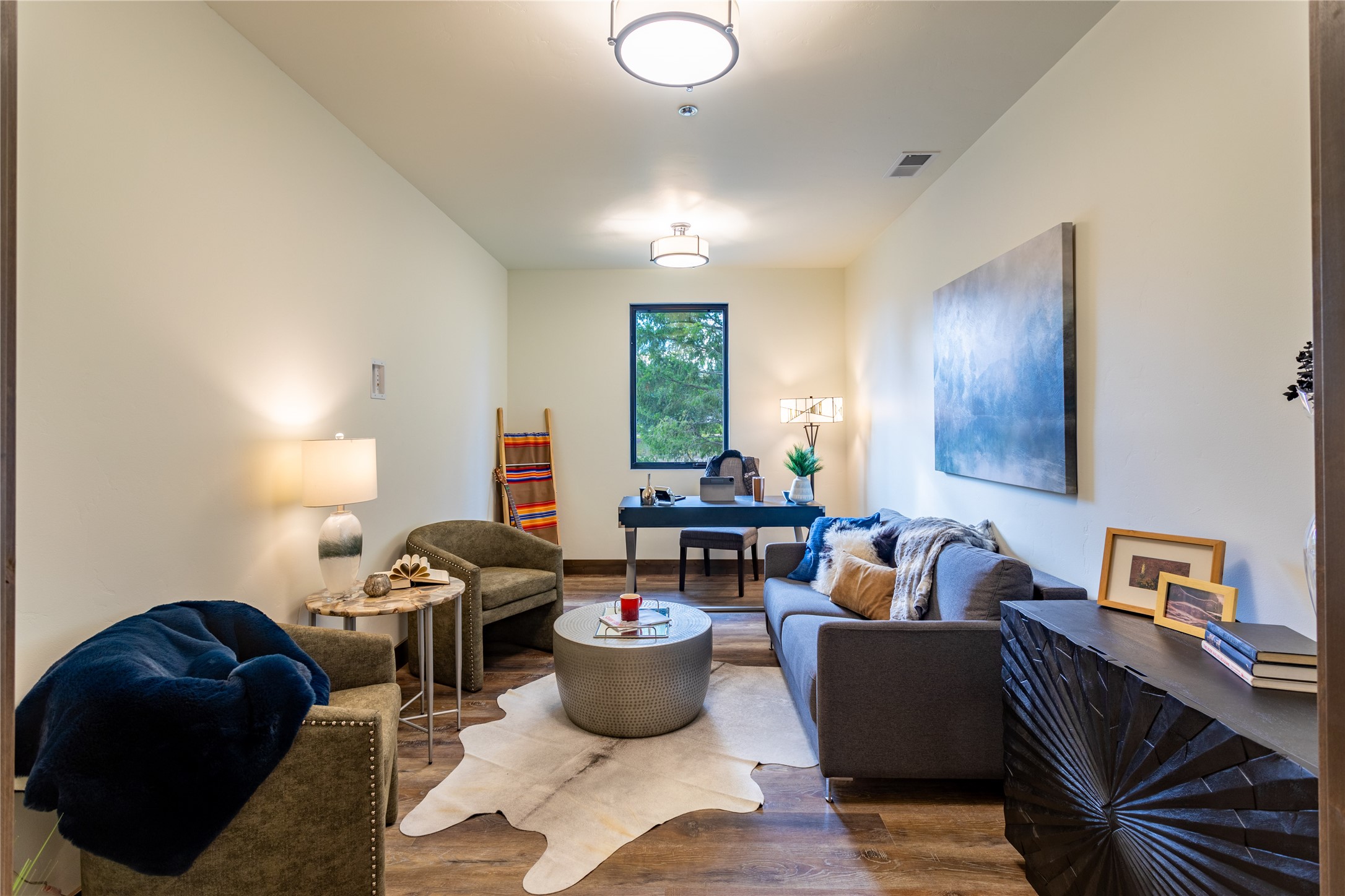 Staged living room with gray sofa, armchairs, hide rug, and a desk beneath the window.