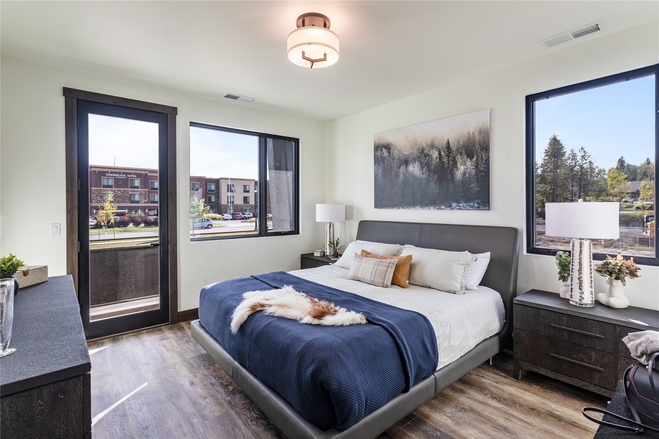 Staged bedroom with a gray upholstered bed, navy blanket, and large windows overlooking nearby buildings and trees.