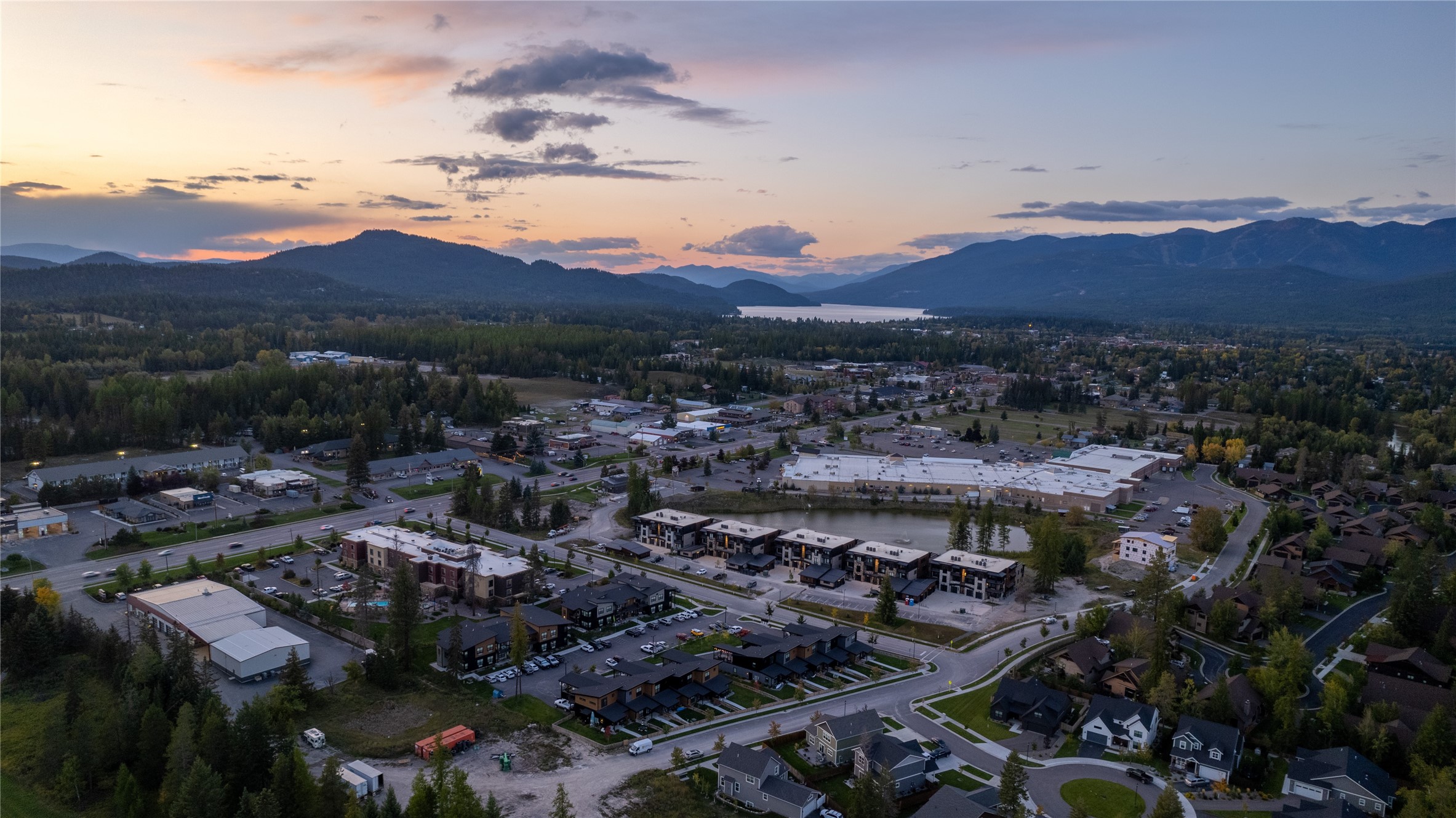 Aerial view of a town, lake, and forested mountains beneath a colorful dusk sky.