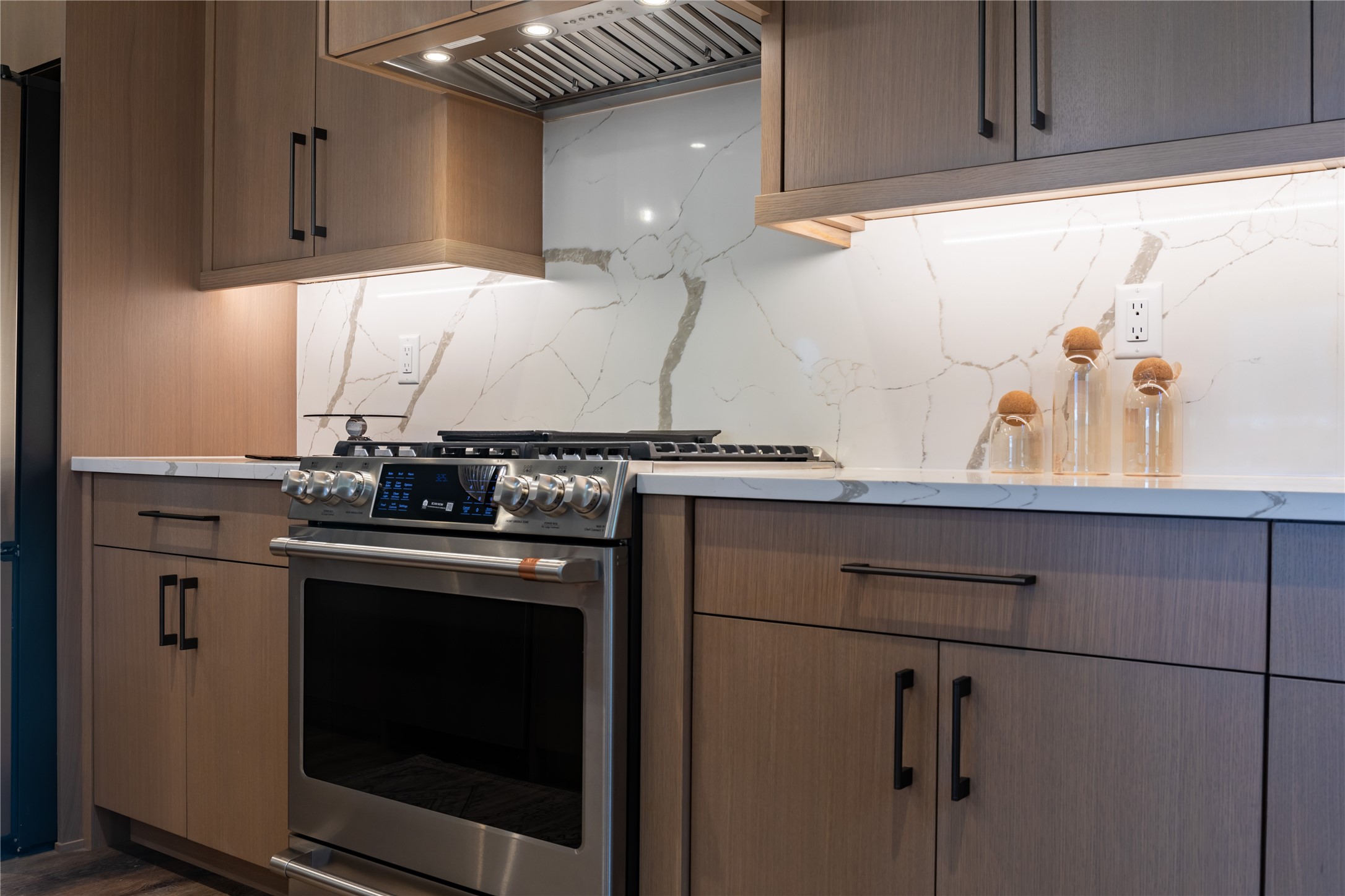 Modern kitchen with a stainless-steel gas range, warm wood cabinets, and a veined stone backsplash.