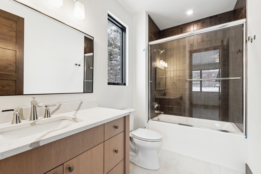 Modern bathroom with a double vanity, glass-enclosed tub, dark tile surround, and snowy view through a narrow window.