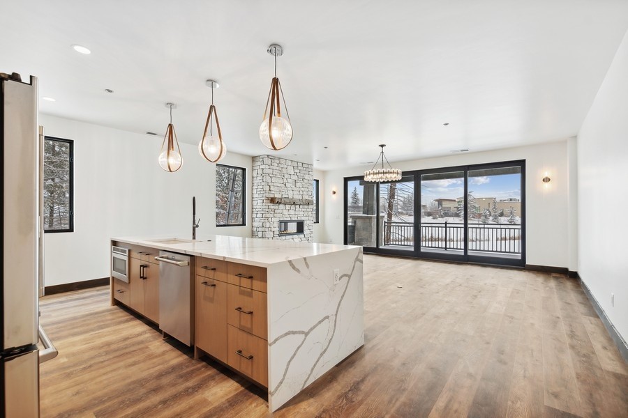 Open-plan kitchen with a marble-look island, stone fireplace, and wide glass doors overlooking a snowy deck.