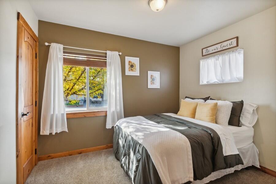 Bedroom with a neatly made bed, white curtains, and a window overlooking yellow autumn leaves.