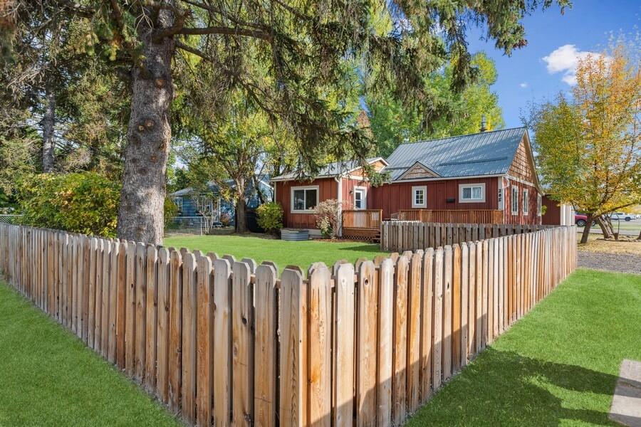 Red single-story home with a metal roof behind a wooden picket fence and large evergreen tree.