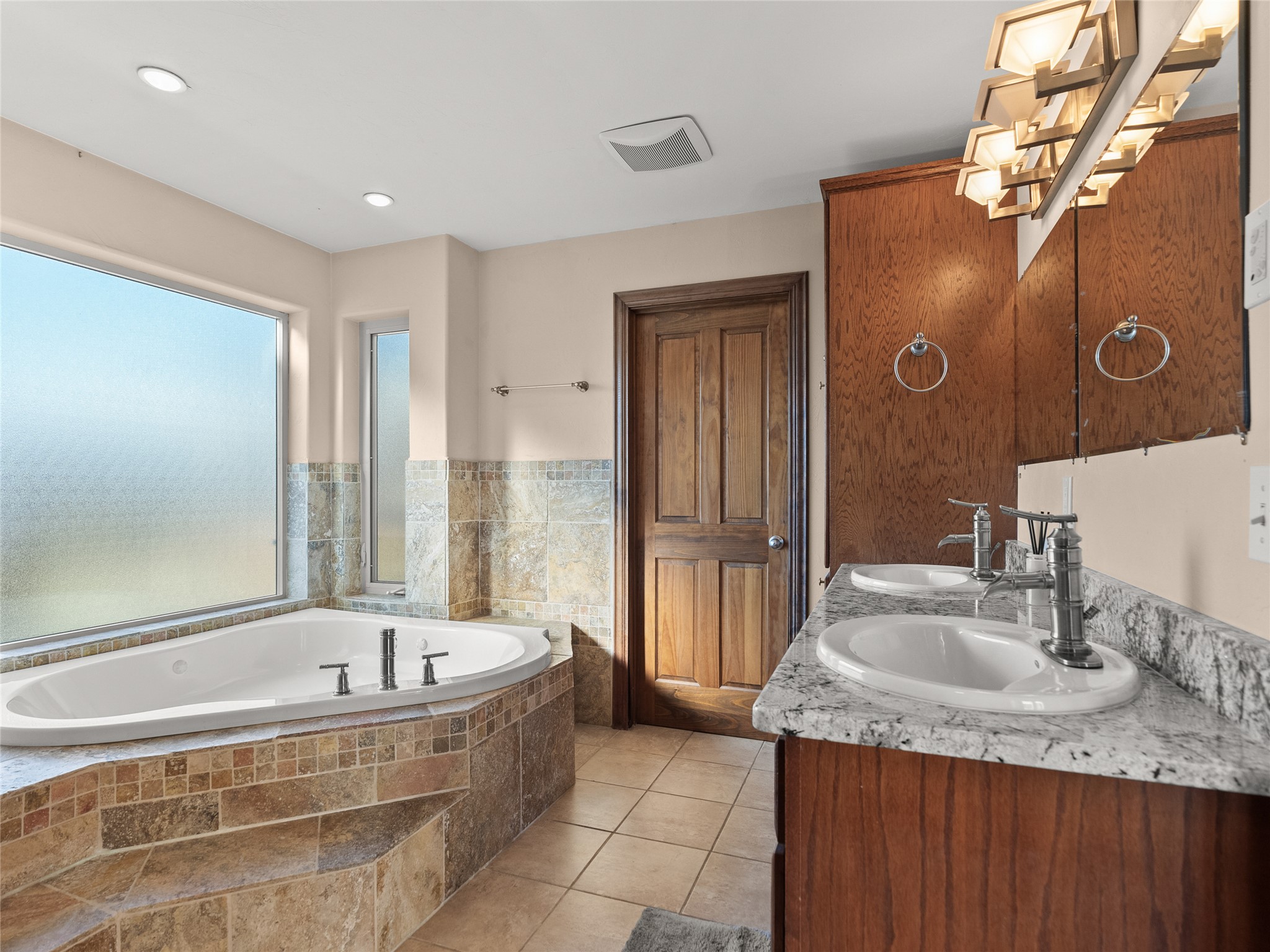Bathroom with a tiled soaking tub beneath frosted windows and a double-sink granite vanity.