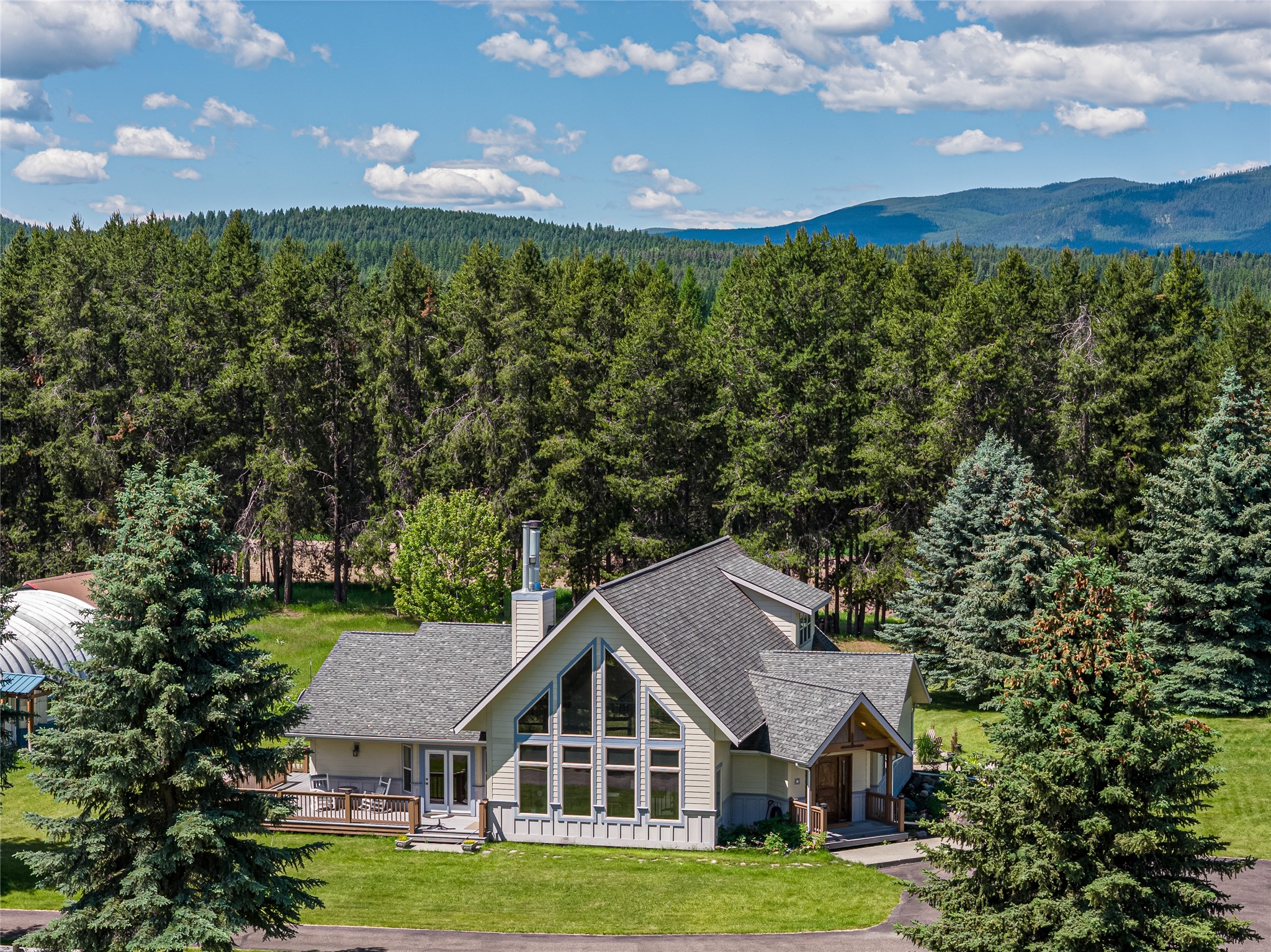 Gray gabled house with large windows, evergreen trees, and forested mountains beyond.