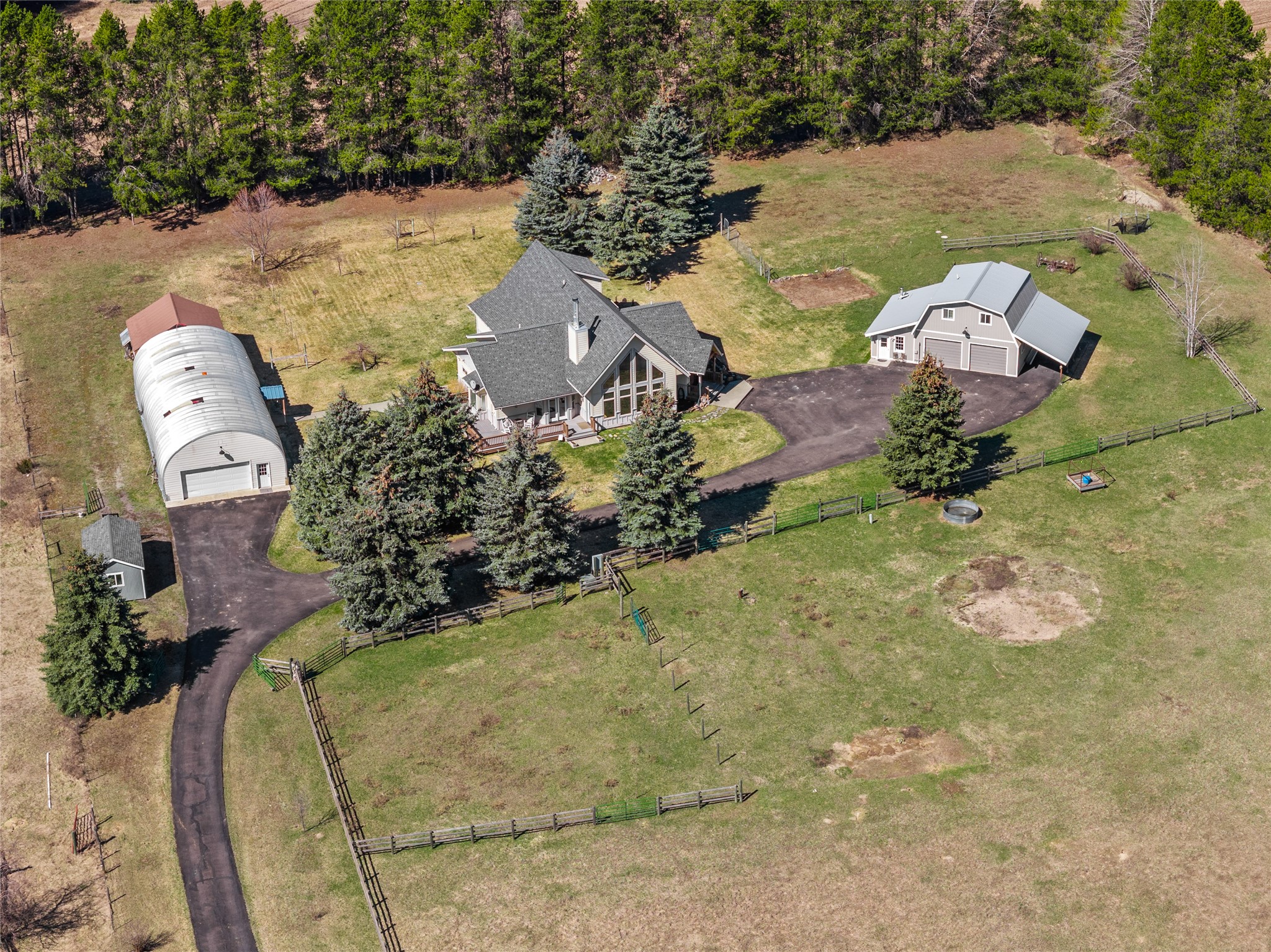 Aerial view of a home, detached barn-style garage, and fenced pasture surrounded by trees.