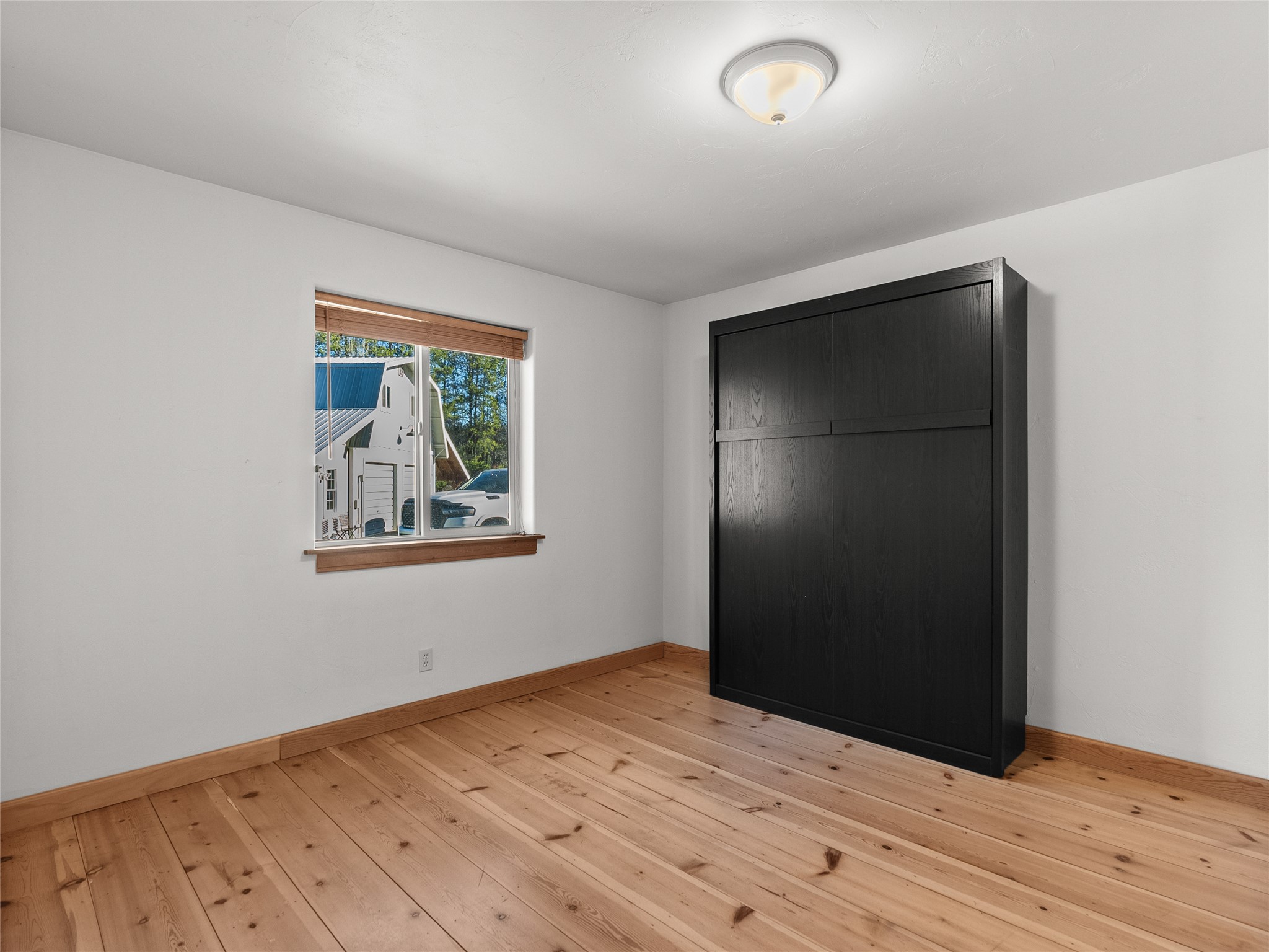 Empty bedroom with natural wood floors, a window with wooden blinds, and a large black wardrobe.
