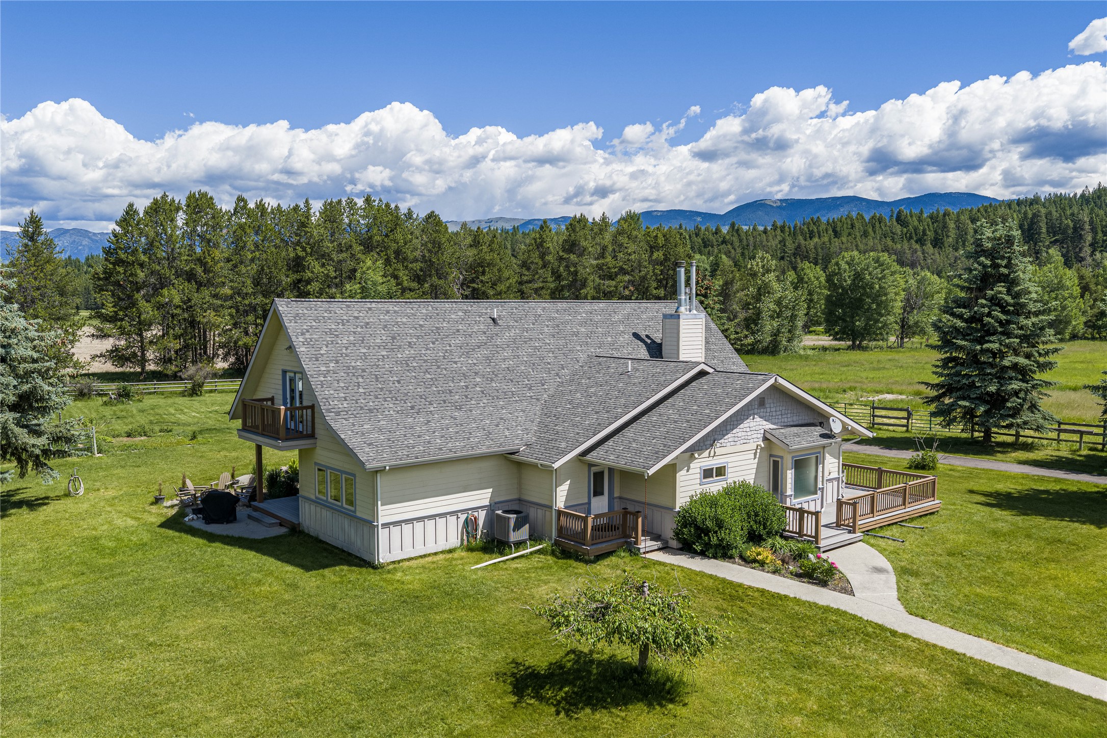 House with multiple decks and a broad lawn, backed by evergreen forest and distant mountains.