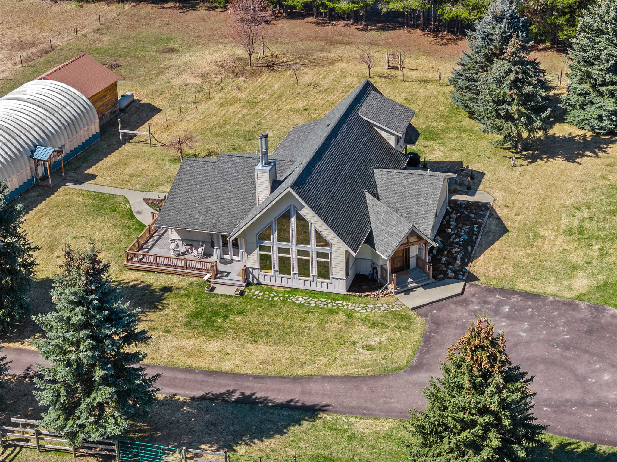 Aerial view of a gray home with tall windows, a wooden deck, evergreen trees, and outbuildings.