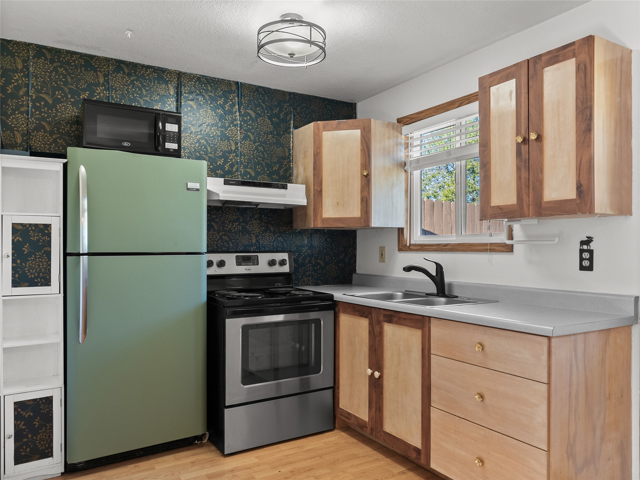 Kitchen with a green refrigerator, floral wallpaper, wood cabinets, and a double-basin sink.