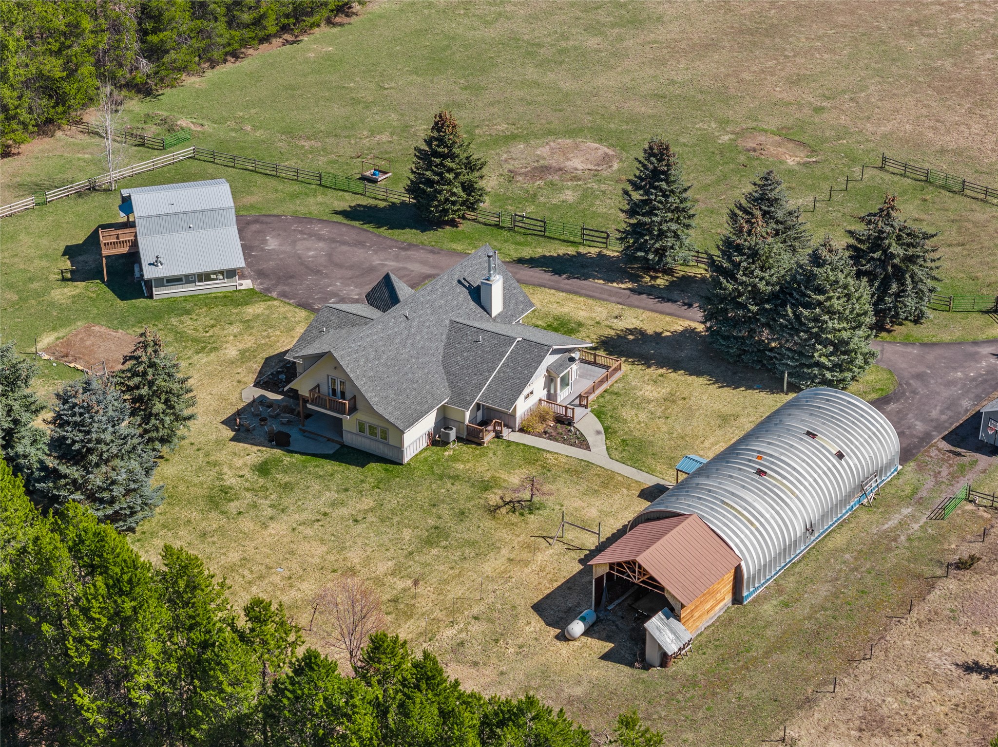 Aerial view of a rural home with a long arched barn, detached outbuilding, and fenced pasture.