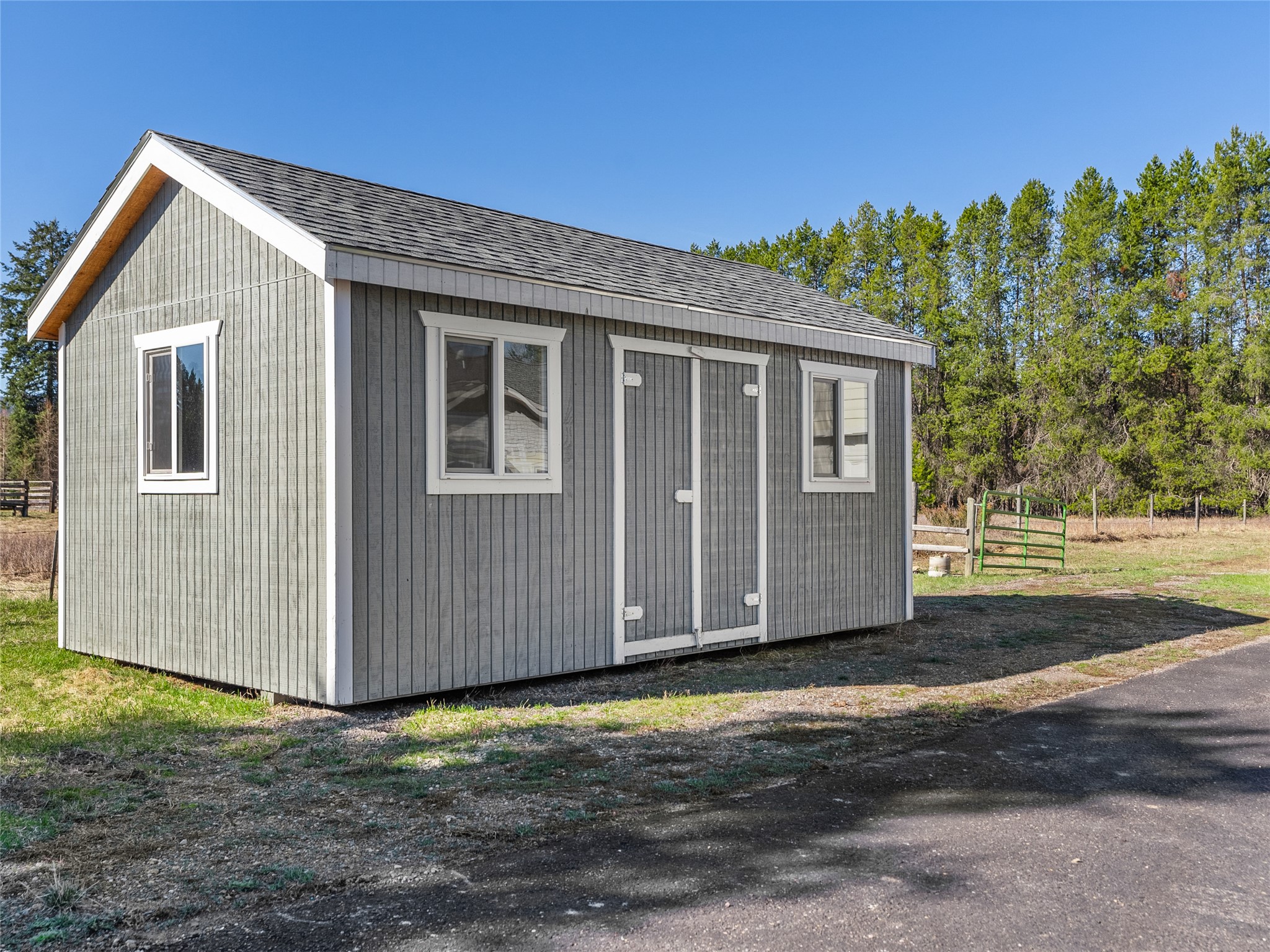 Gray storage shed with white trim, two windows, and double doors beside a gravel drive.