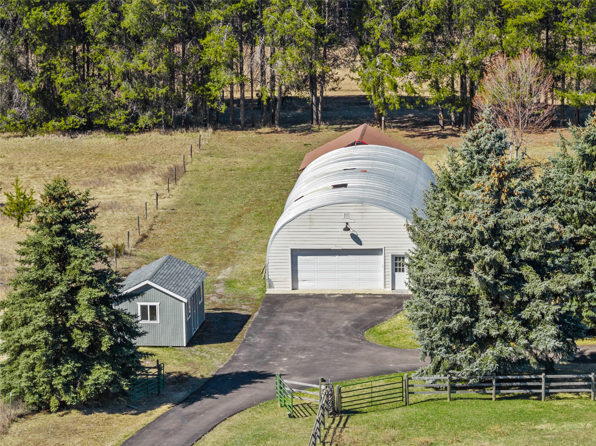 Aerial view of an arched-roof outbuilding and small shed amid trees and fenced grounds.