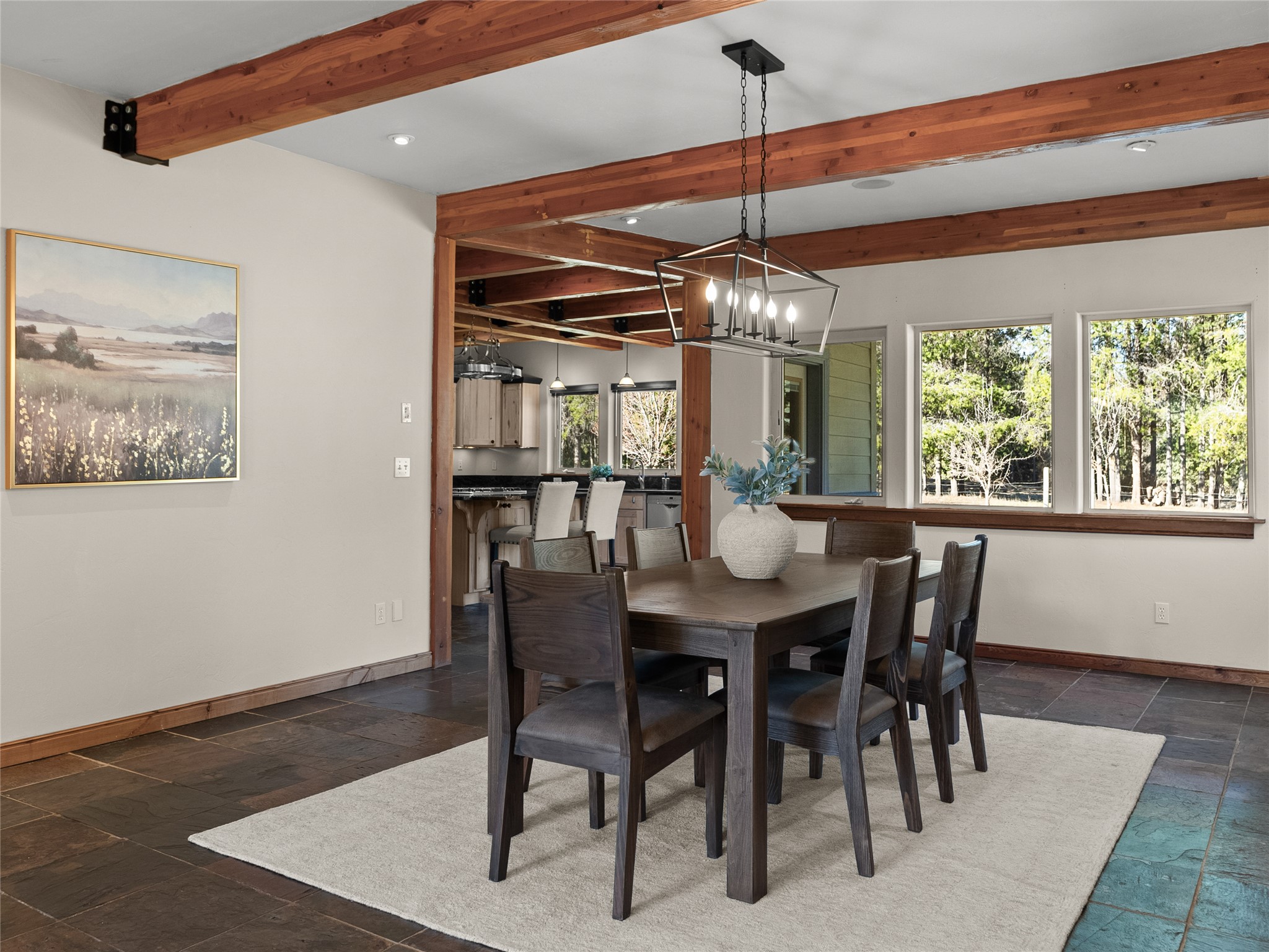 Dining room with exposed timber beams, a dark wood table, and large windows overlooking trees.