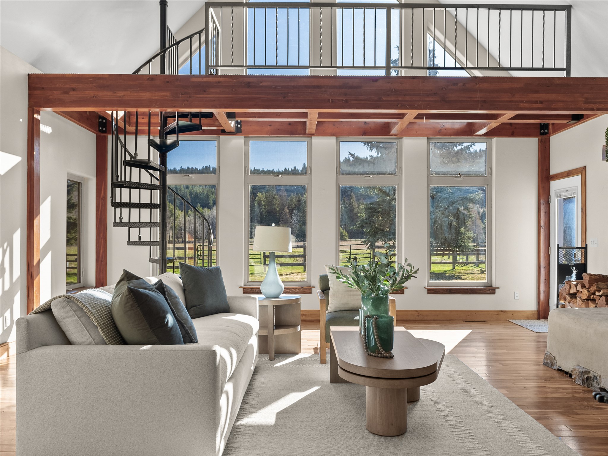 Furnished living room with a spiral staircase, timber loft, and tall windows overlooking trees.