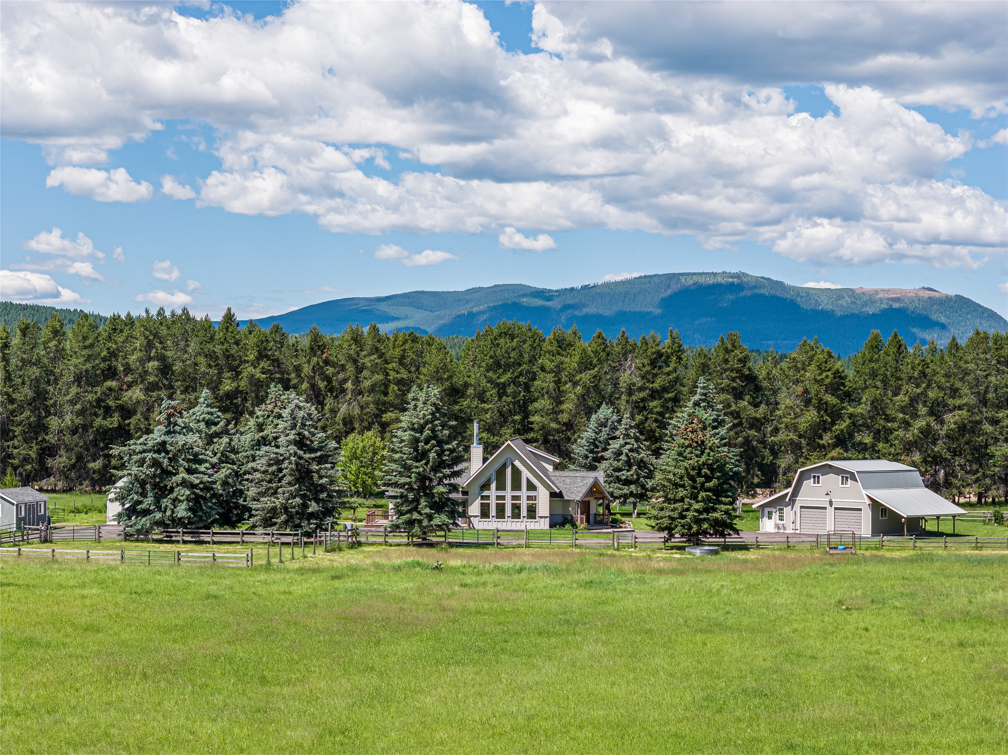 House and barn-style garage across a fenced green field, backed by evergreens and forested mountains.