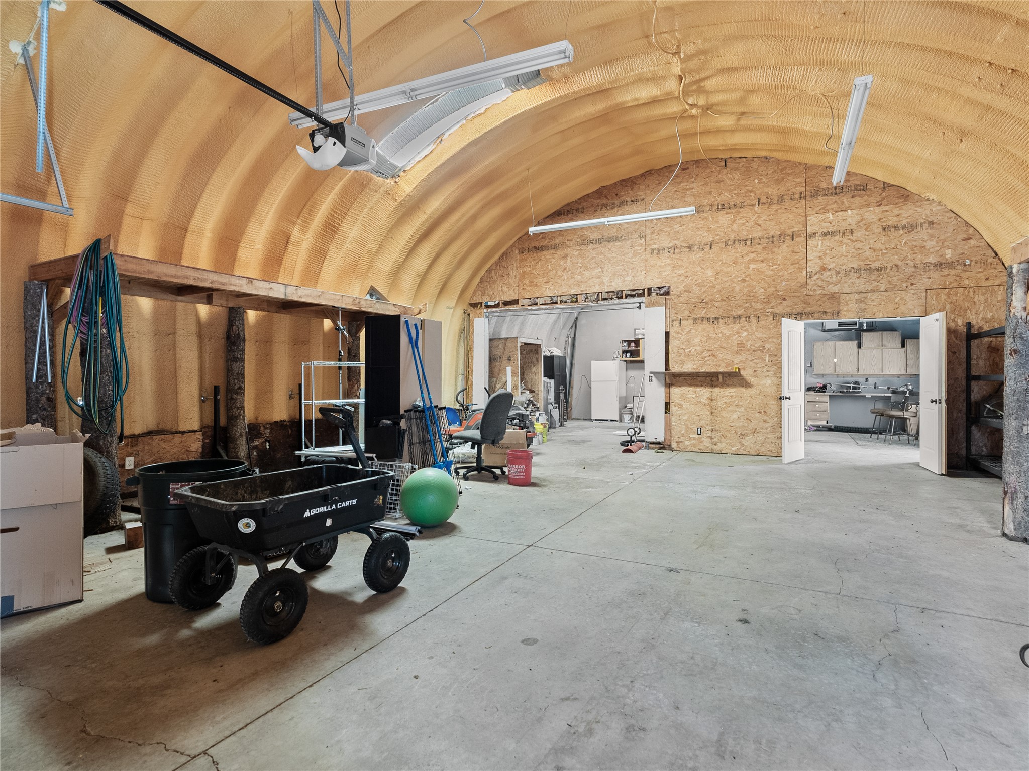 Workshop with an arched insulated ceiling, concrete floor, equipment carts, and a kitchen through open double doors.