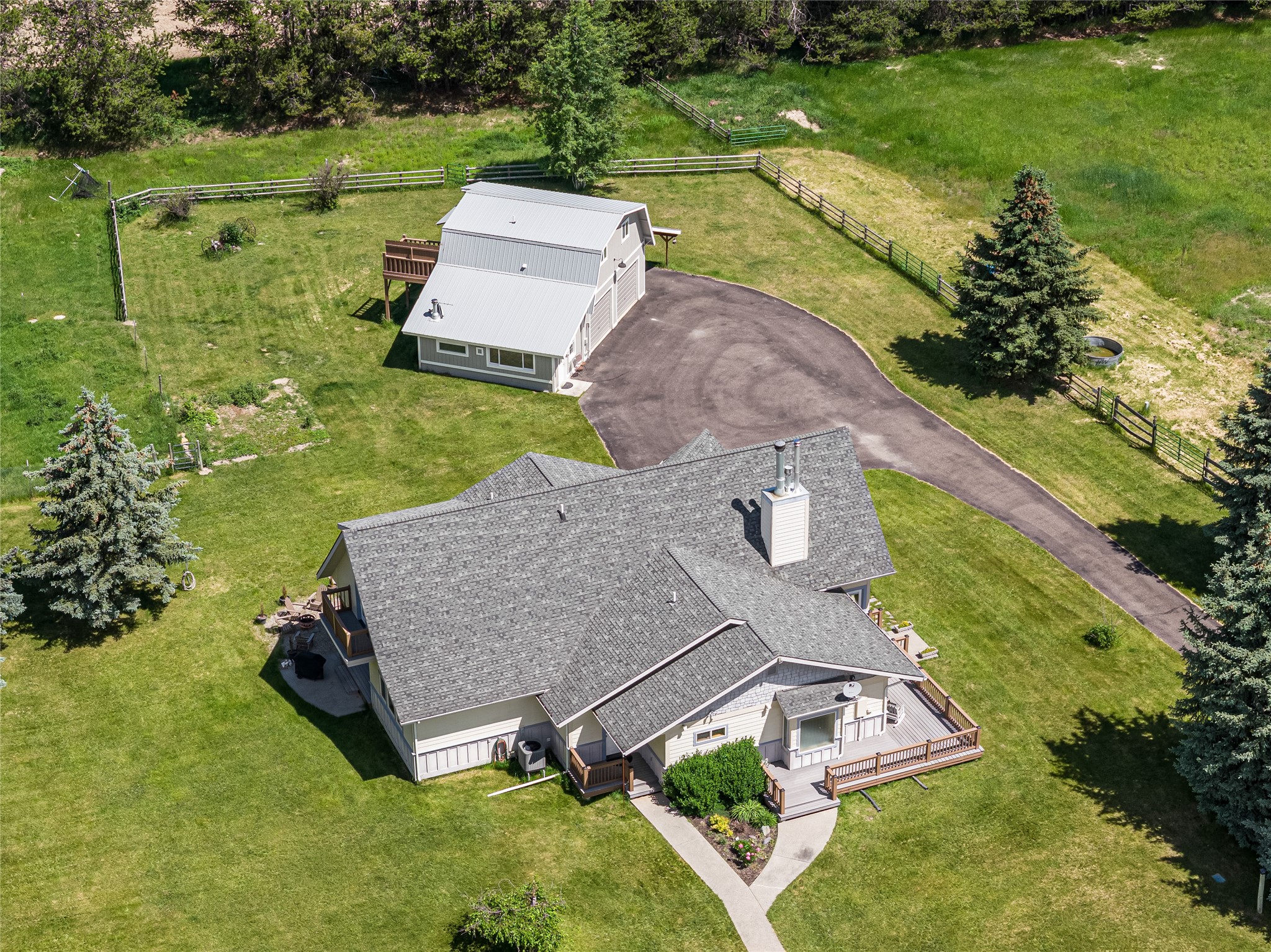 Aerial view of a home and outbuilding, with a curved driveway, fenced lawn, and mature evergreens.