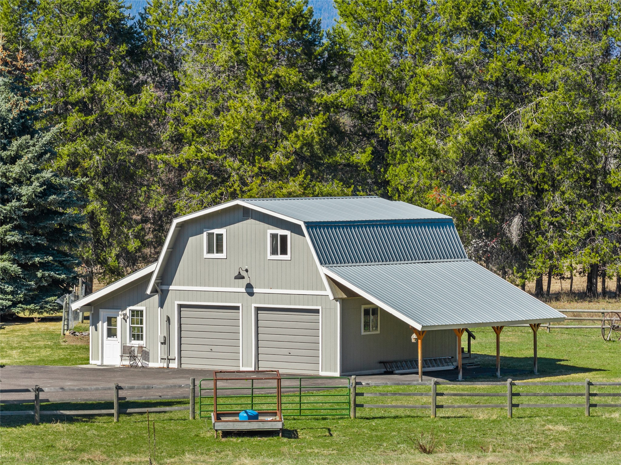 Gray barn-style garage with two doors and a large covered carport behind a fenced yard.