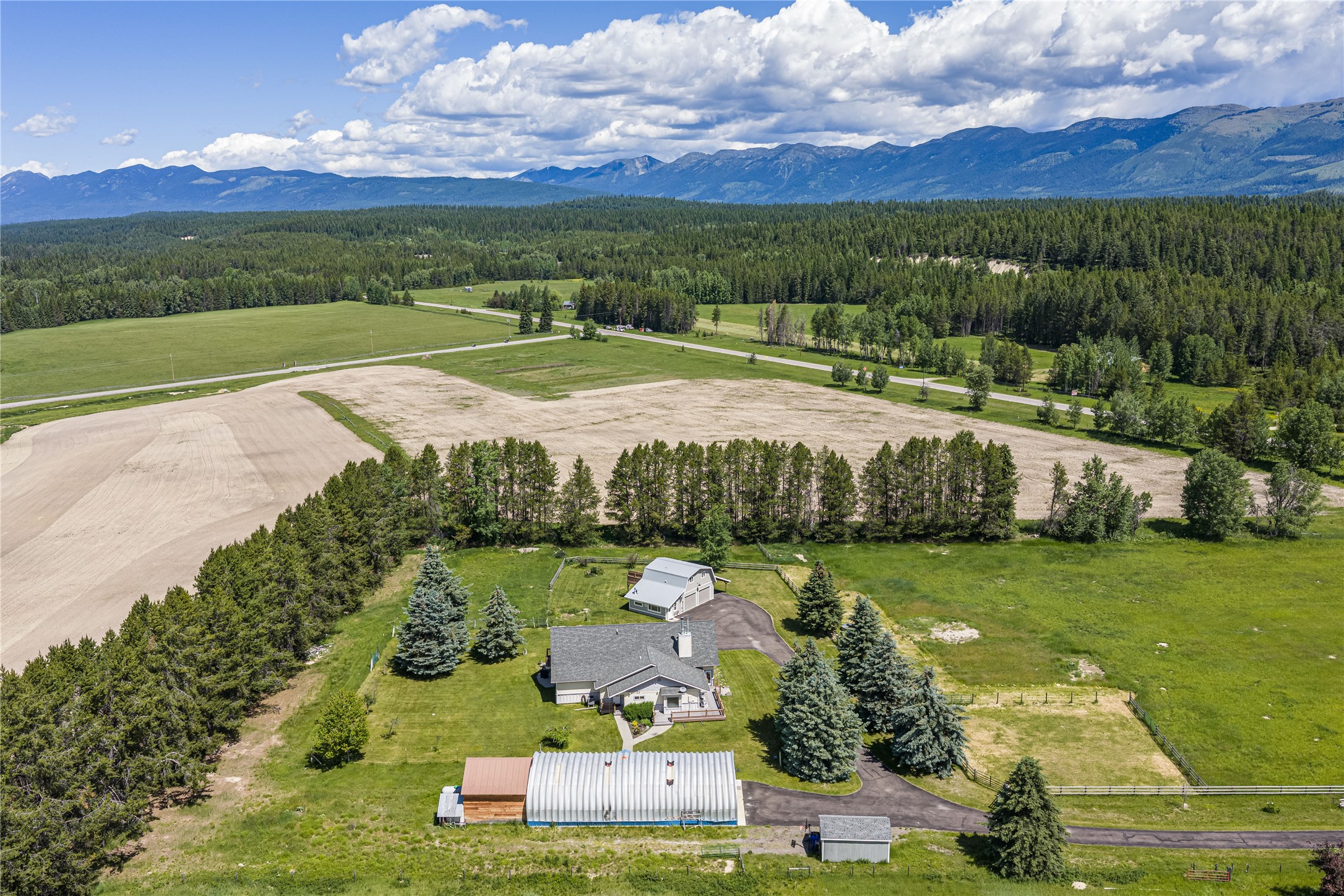 Aerial view of a rural home and outbuildings amid fields, forest, and distant mountains.